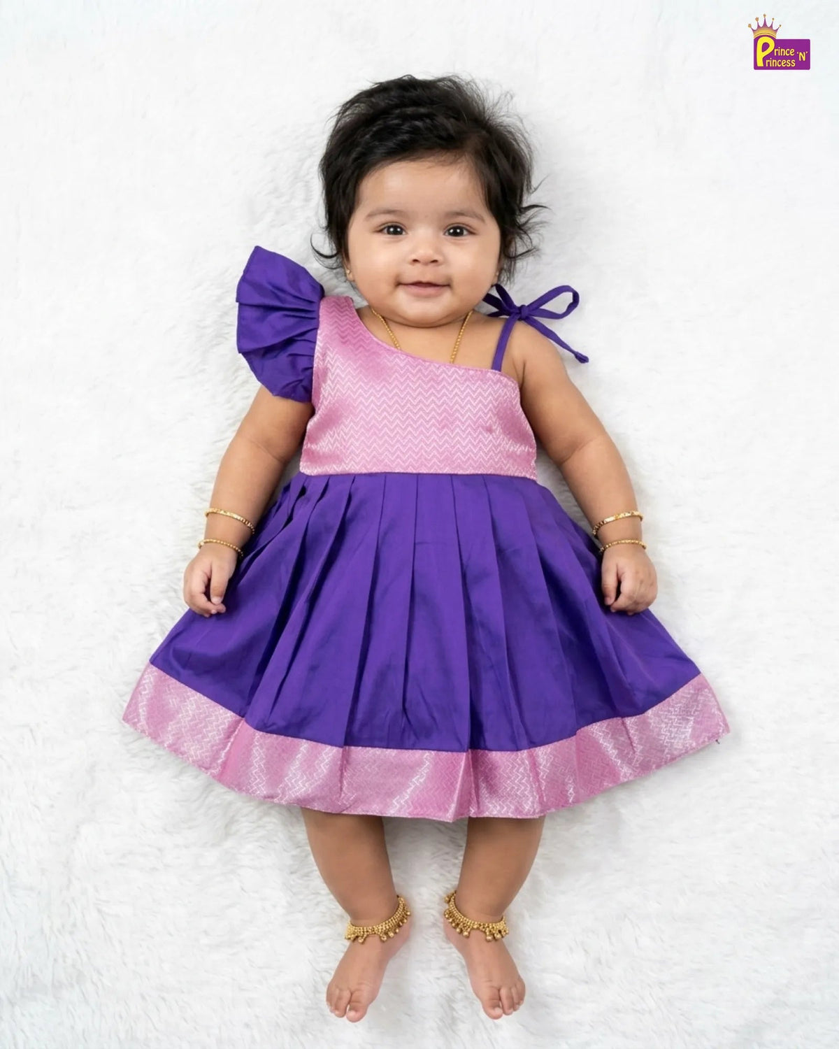 A baby girl wearing a purple and pink dress, lying on a white textured blanket. The dress has a pink patterned bodice, a purple skirt, and a purple bow on the shoulder.