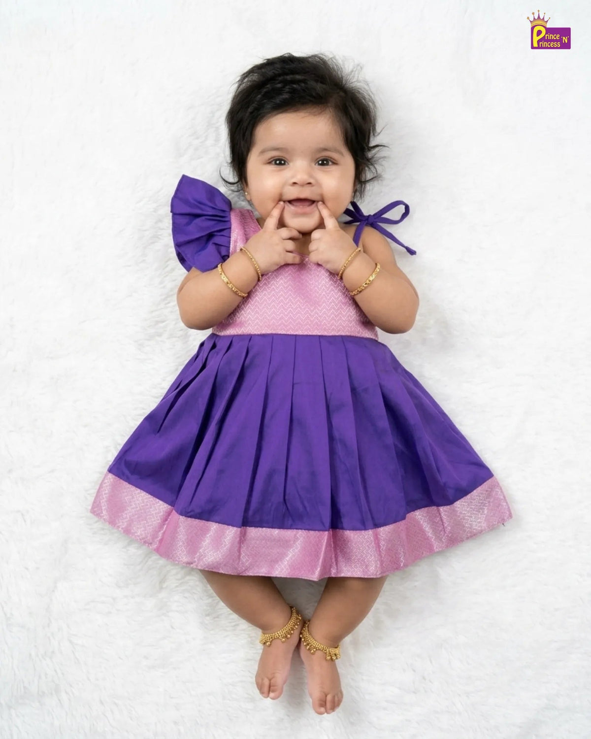 A baby girl lying on a white textured blanket, wearing a purple and pink dress with a pleated skirt, a pink polka-dot bodice, and a purple bow on her shoulder.