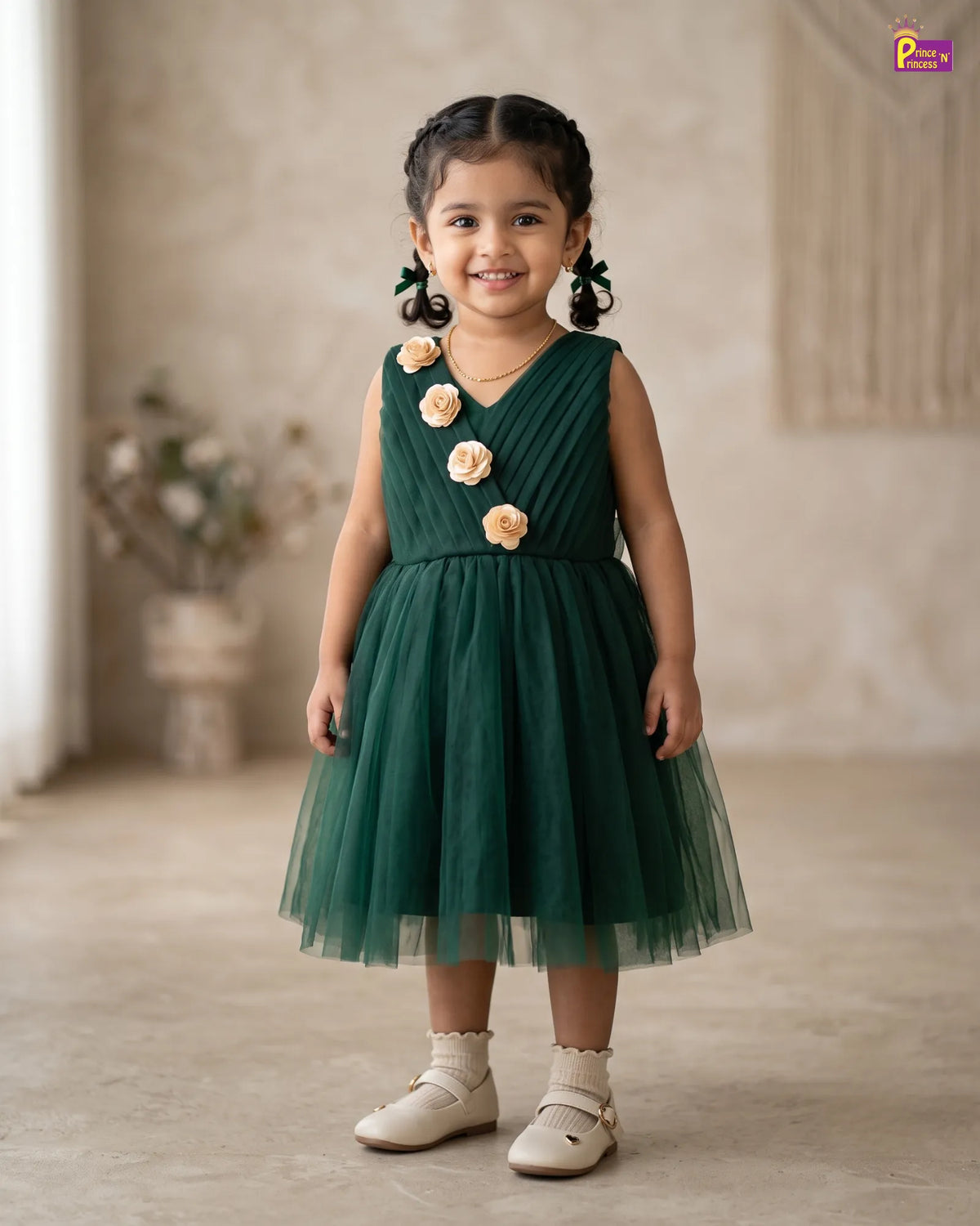 A young girl with dark hair in pigtails stands wearing a sleeveless dark green tulle dress decorated with three large cream-colored fabric flowers on the bodice. She is wearing cream-colored shoes and standing in a softly lit indoor room with a textured beige wall and floor.