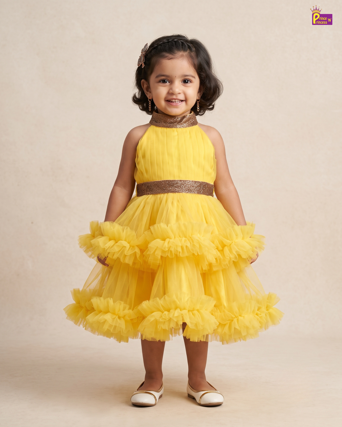 A young girl with dark curly hair stands wearing a bright yellow sleeveless party dress with a ruffled tulle skirt and a metallic gold glittery waistband, set against a plain beige studio background.