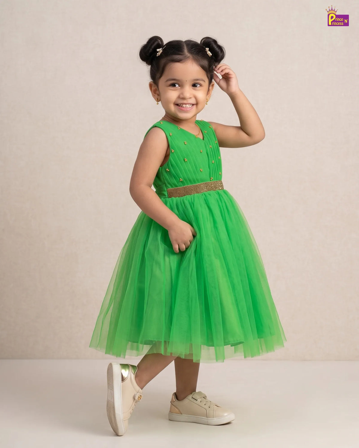 A young girl with her hair in two buns poses in a sleeveless bright green tulle dress with a metallic gold belt, standing against a neutral beige background.