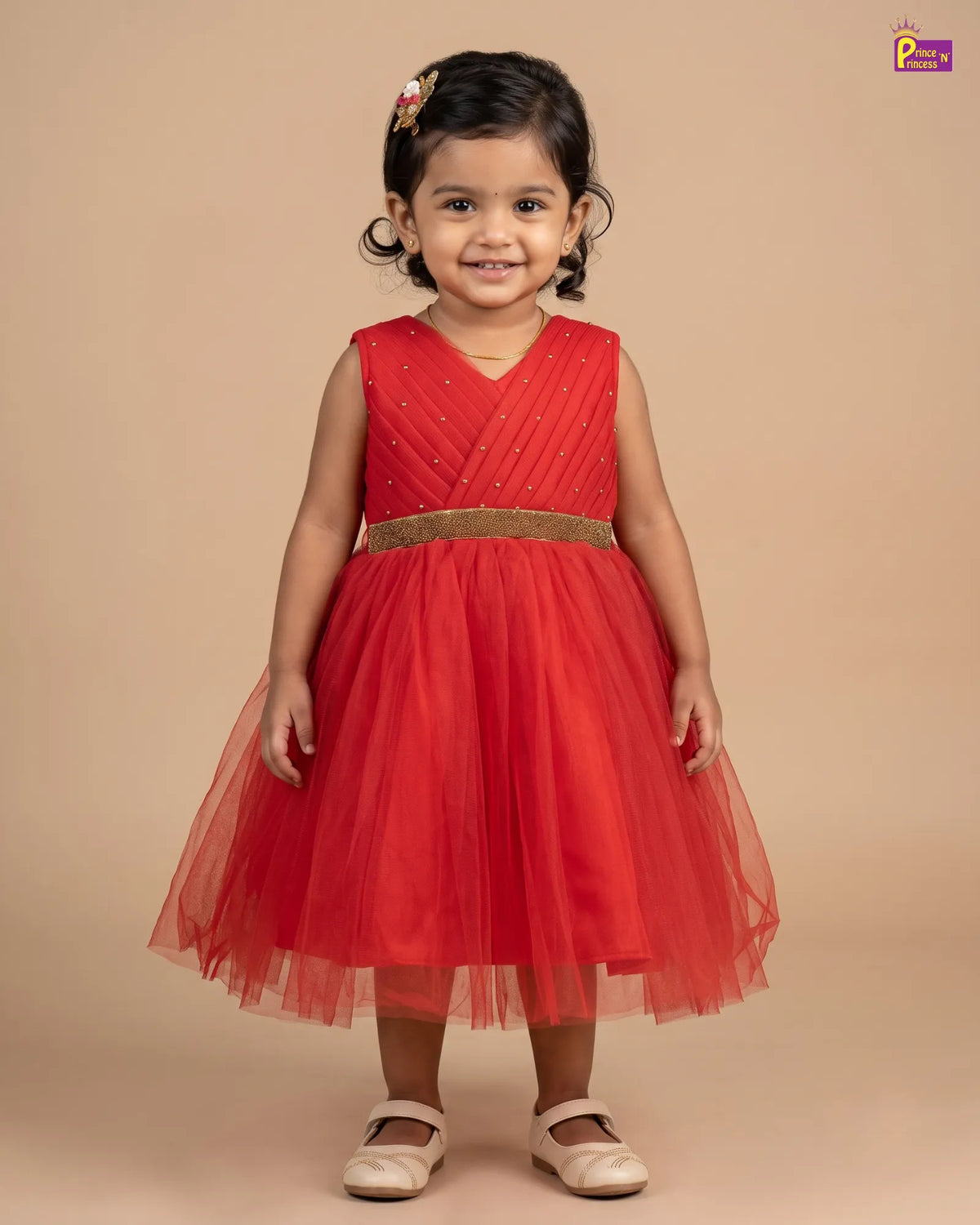 A young girl with dark hair wearing a sleeveless red party dress with a gold waistband and a full tulle skirt, standing against a solid beige background.