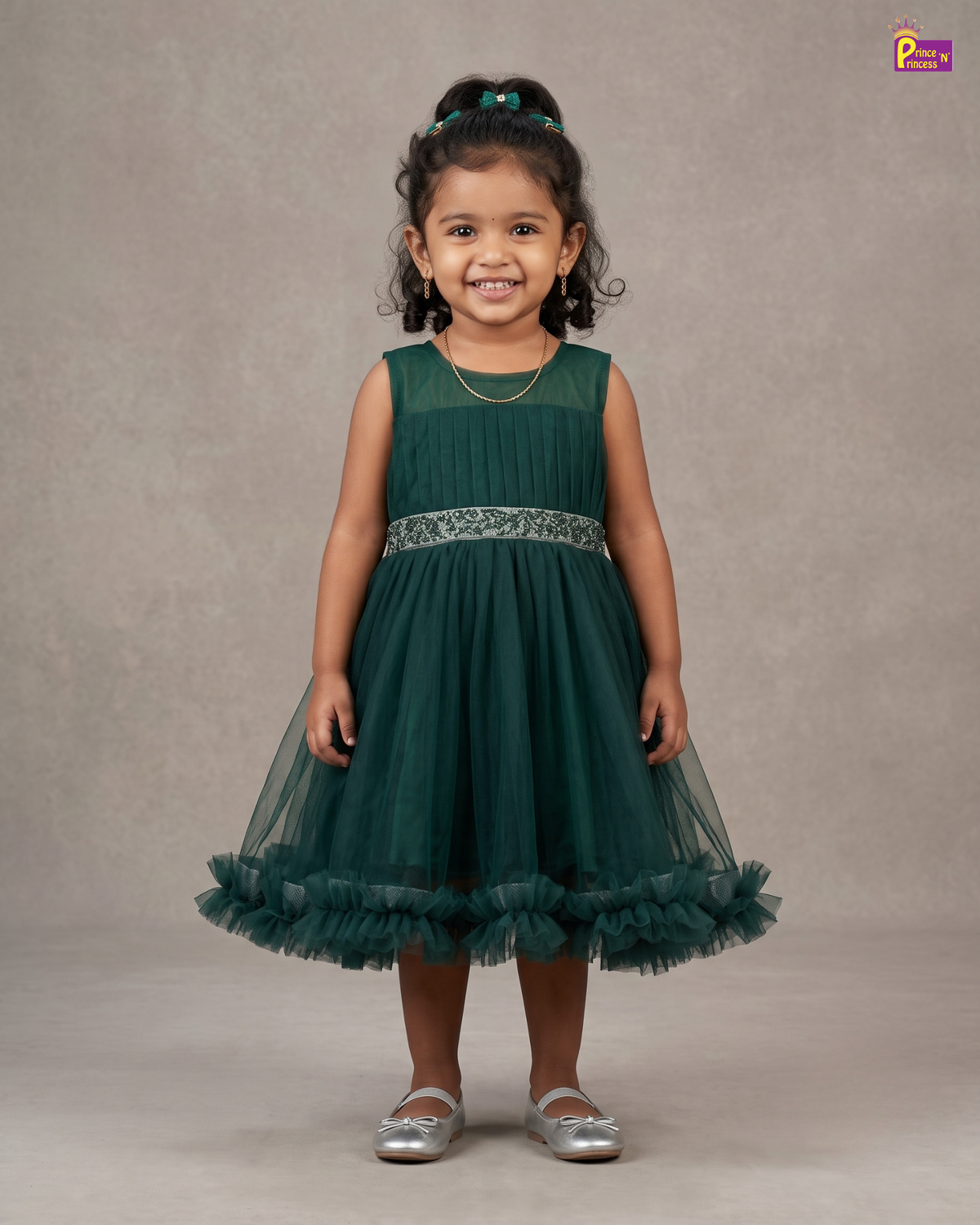 A young girl with curly dark hair wearing a sleeveless dark green tulle flower girl dress with a ruffled hem and a silver patterned waistband, standing against a neutral gray studio backdrop.
