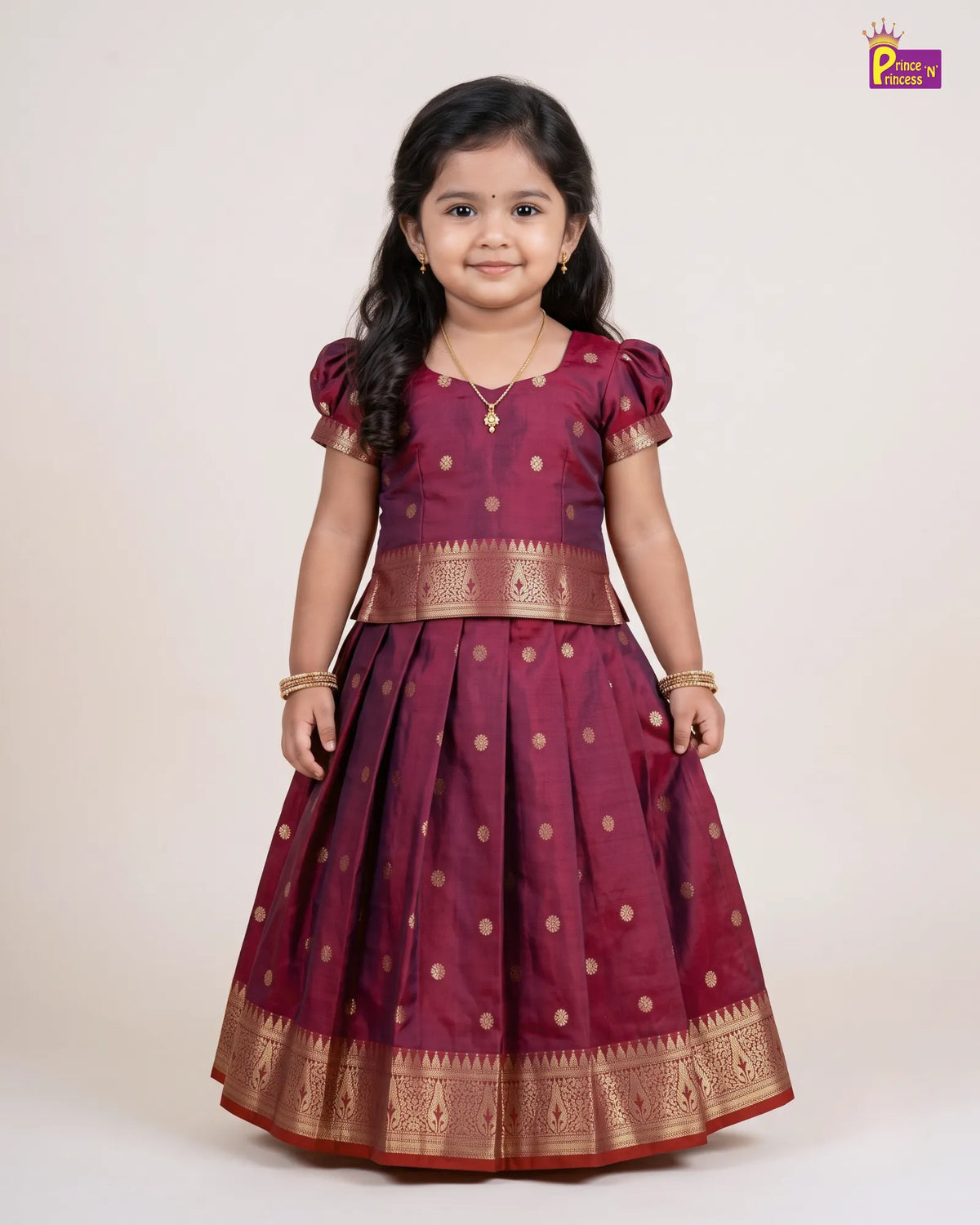 A young girl modeling a traditional Indian dress, featuring a deep maroon body with gold polka dots and a wide gold zari border, paired with a matching gold-embellished waistband and blouse. The image is set against a plain light beige background with a 'Photo by Princi' watermark in the top right corner.