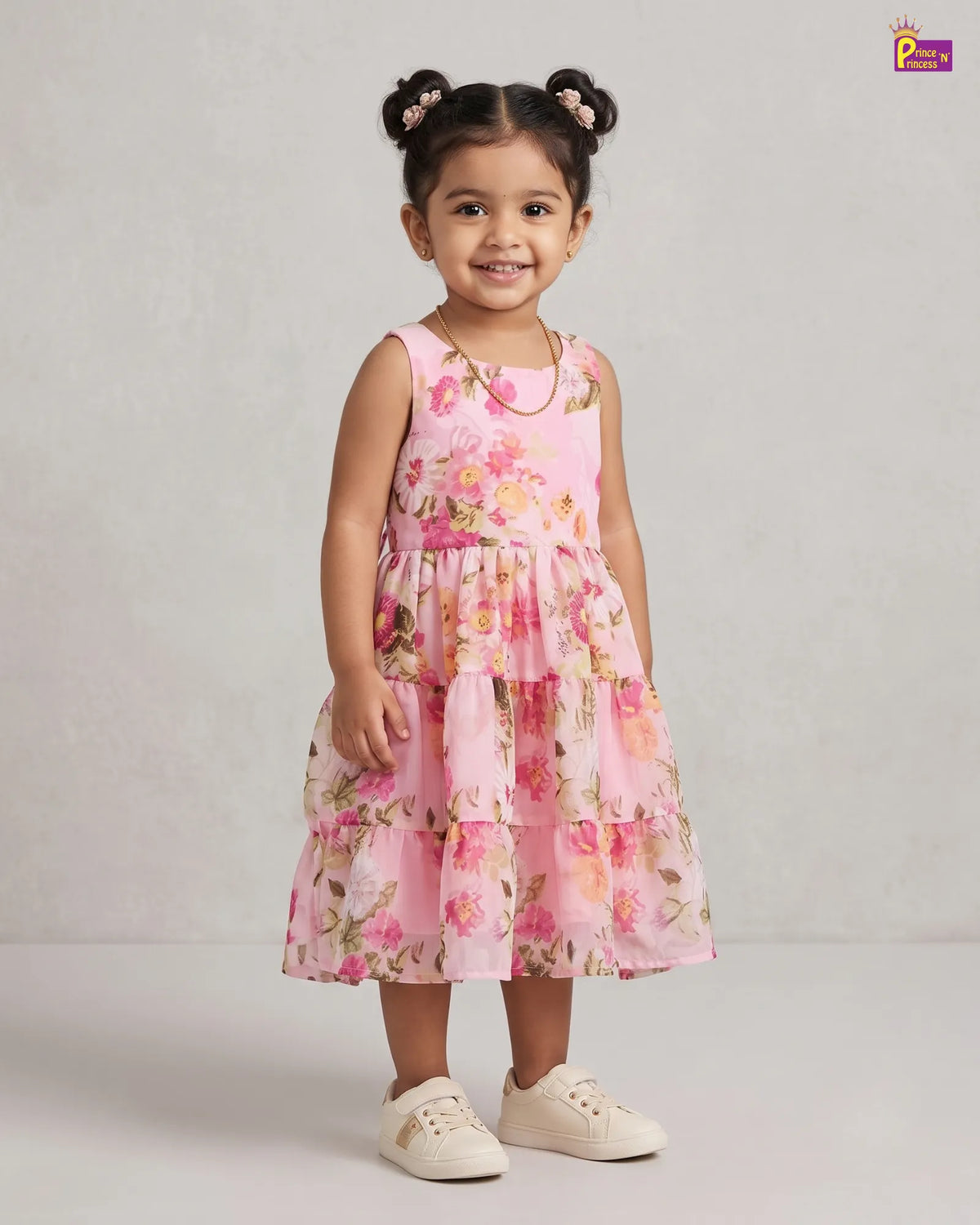 A young girl with her hair in two buns stands wearing a tiered pink floral dress with white sneakers against a light gray studio background.