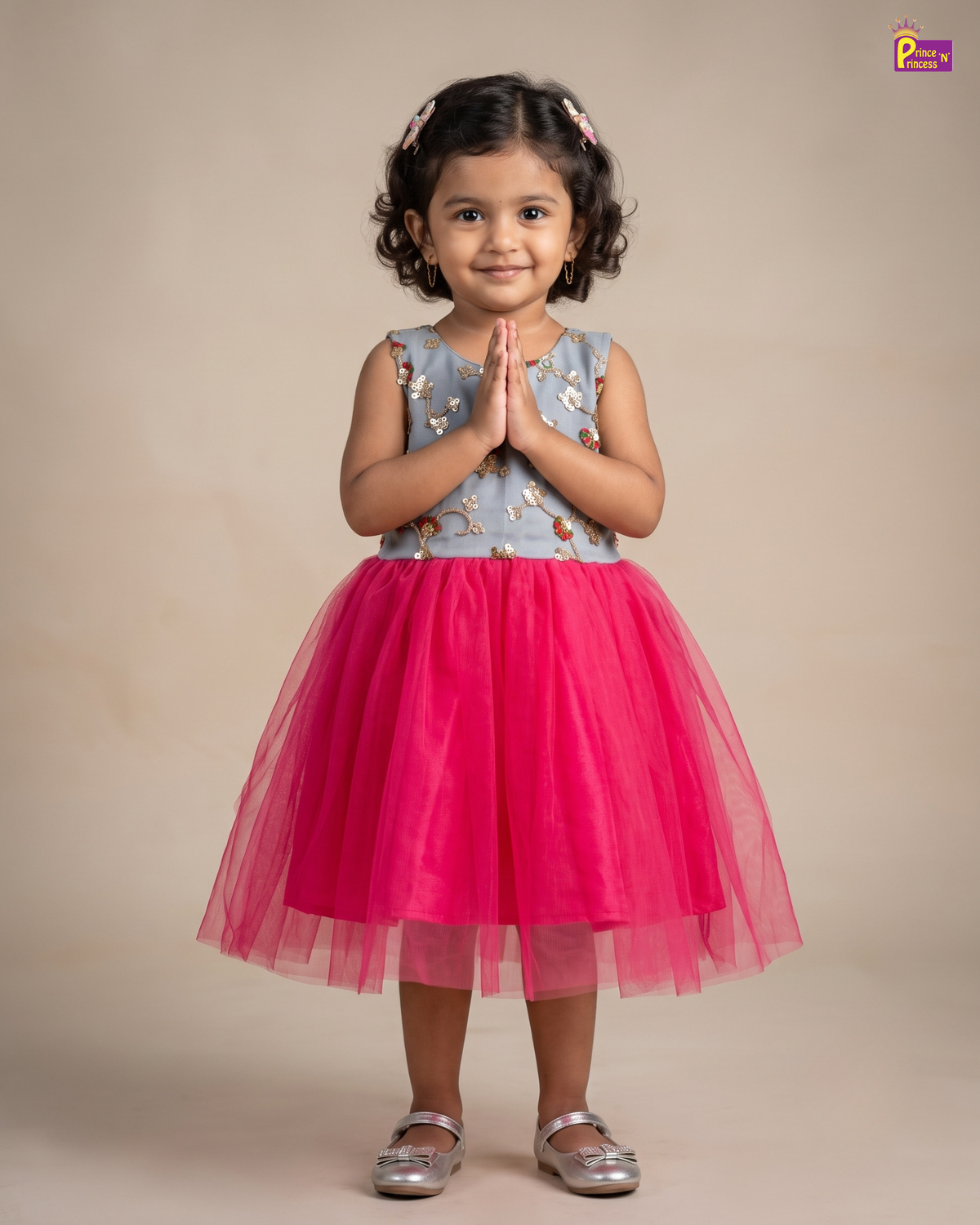 A young girl with curly dark hair wearing a sleeveless light blue floral top and a bright pink tulle skirt, standing with hands pressed together in a studio setting.