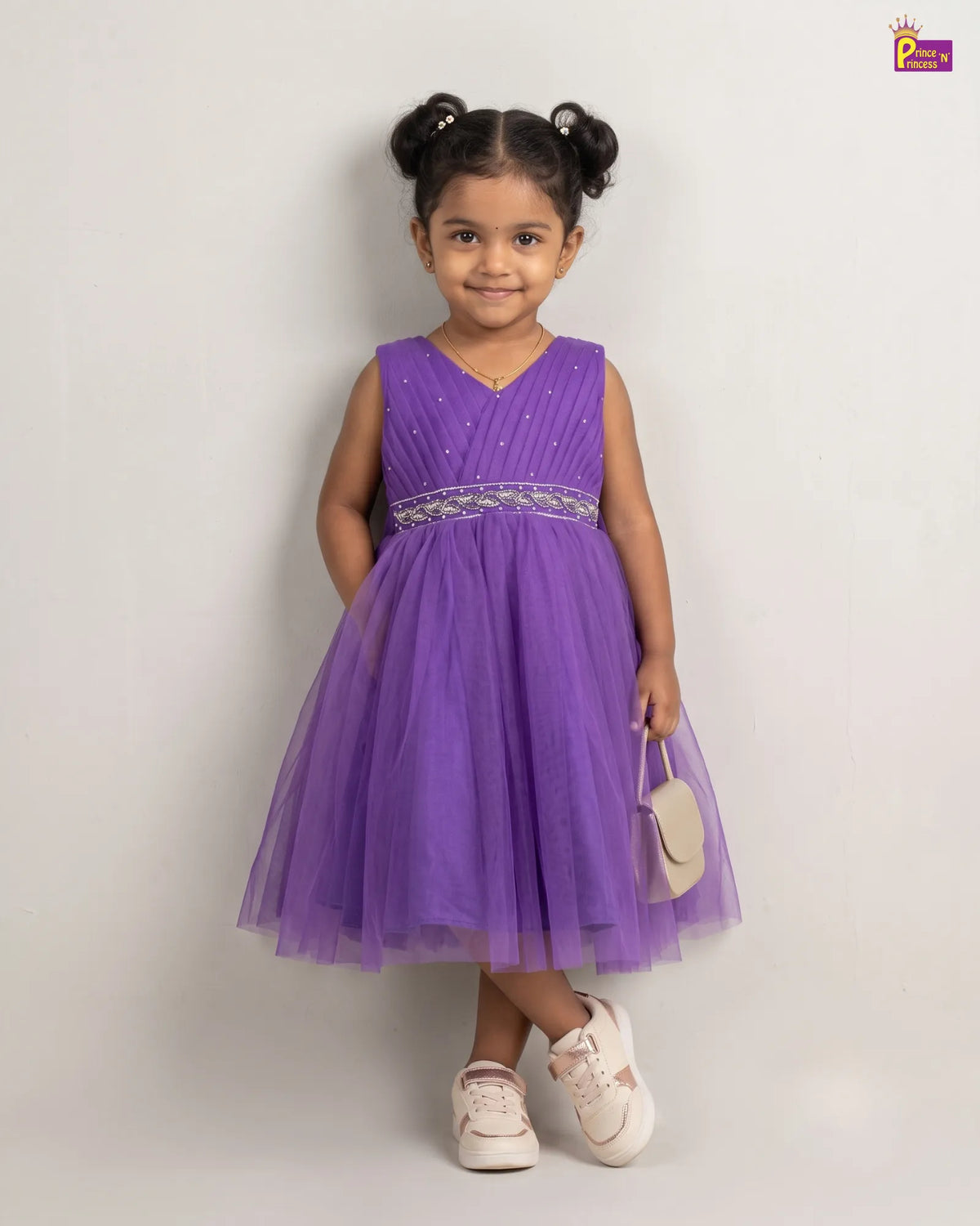 A young girl with her hair in two buns stands against a light gray studio background wearing a sleeveless purple tulle dress with a sparkly silver waistband and holding a small beige handbag.