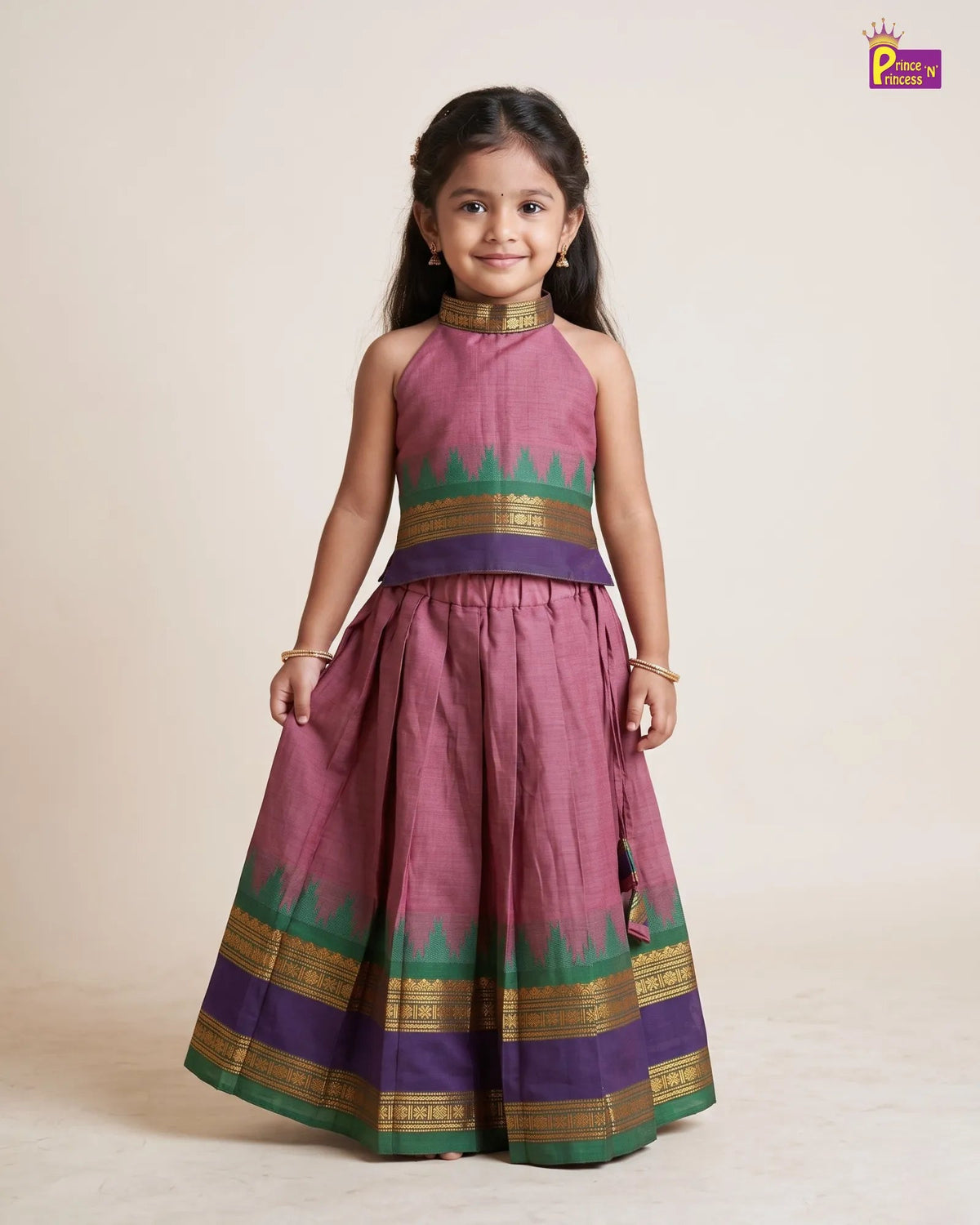 A young girl modeling a traditional Indian salwar kameez dress in dusty pink with green and purple borders and gold zari detailing, set against a plain beige studio background.