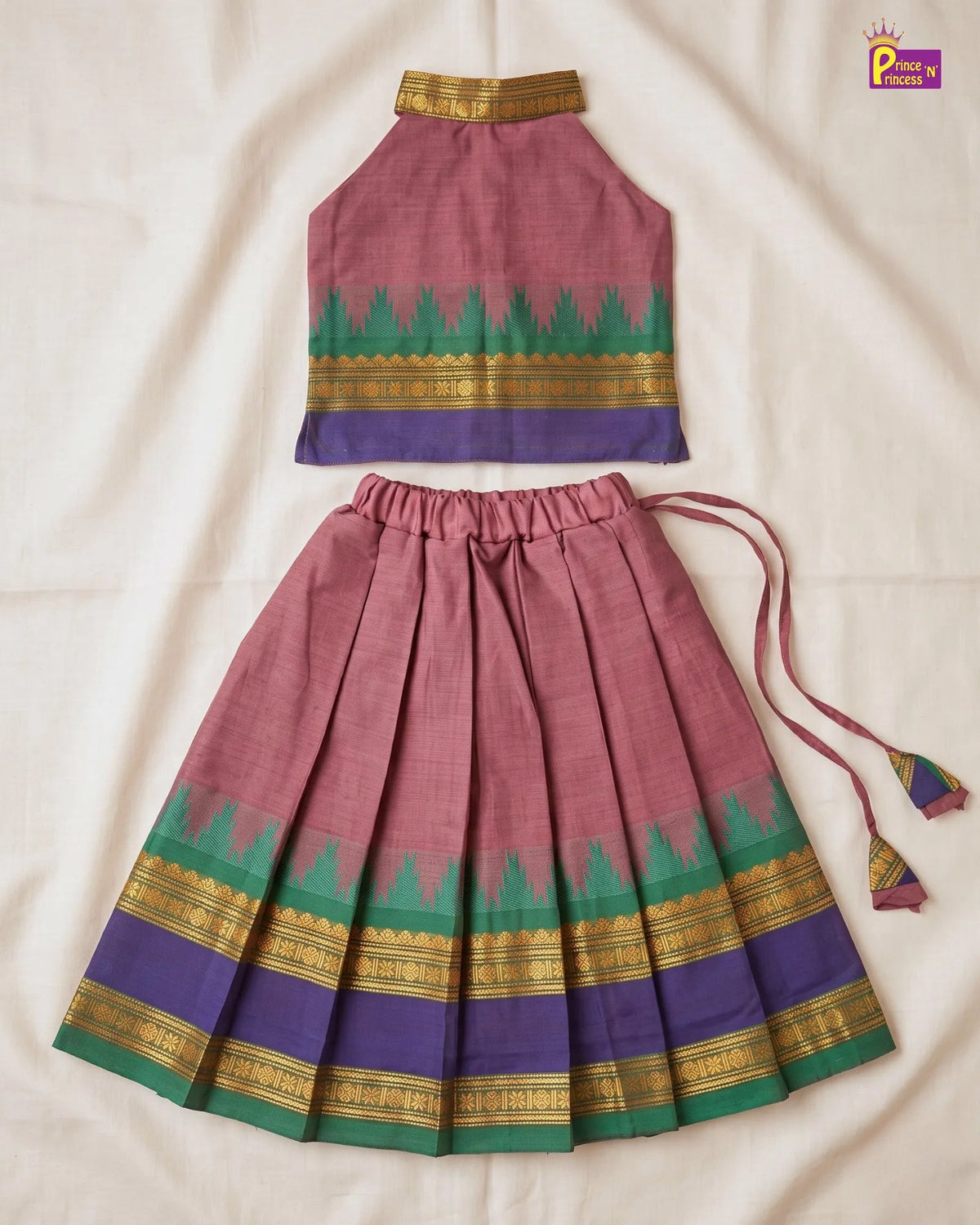 A traditional Indian two-piece outfit made of dusty pink cotton fabric, featuring a sleeveless halter top and a pleated skirt with green, purple, and gold patterned borders, laid flat on a cream-colored fabric background.