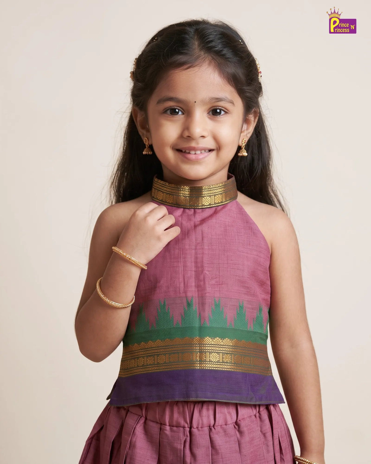 A young girl modeling a traditional Indian salwar kameez outfit. The top is a dusty rose pink with a green and gold zari border, paired with a matching pleated skirt.