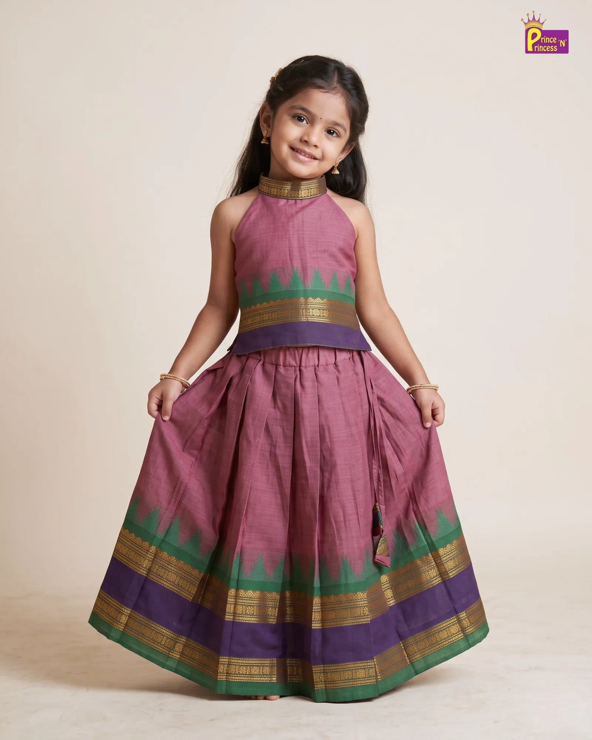 A young girl modeling a sleeveless, high-neck traditional Indian dress with a pleated skirt. The dress features a dusty rose body with green and gold borders, a purple waistband, and a gold embroidered collar.