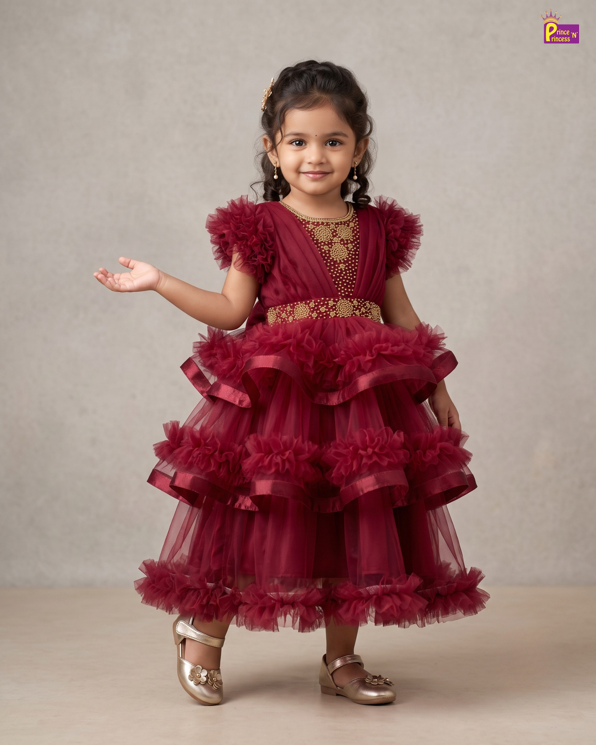 A young girl with braided hair wearing a burgundy tiered tulle party dress with gold embroidery and metallic gold shoes, standing against a neutral beige studio backdrop.