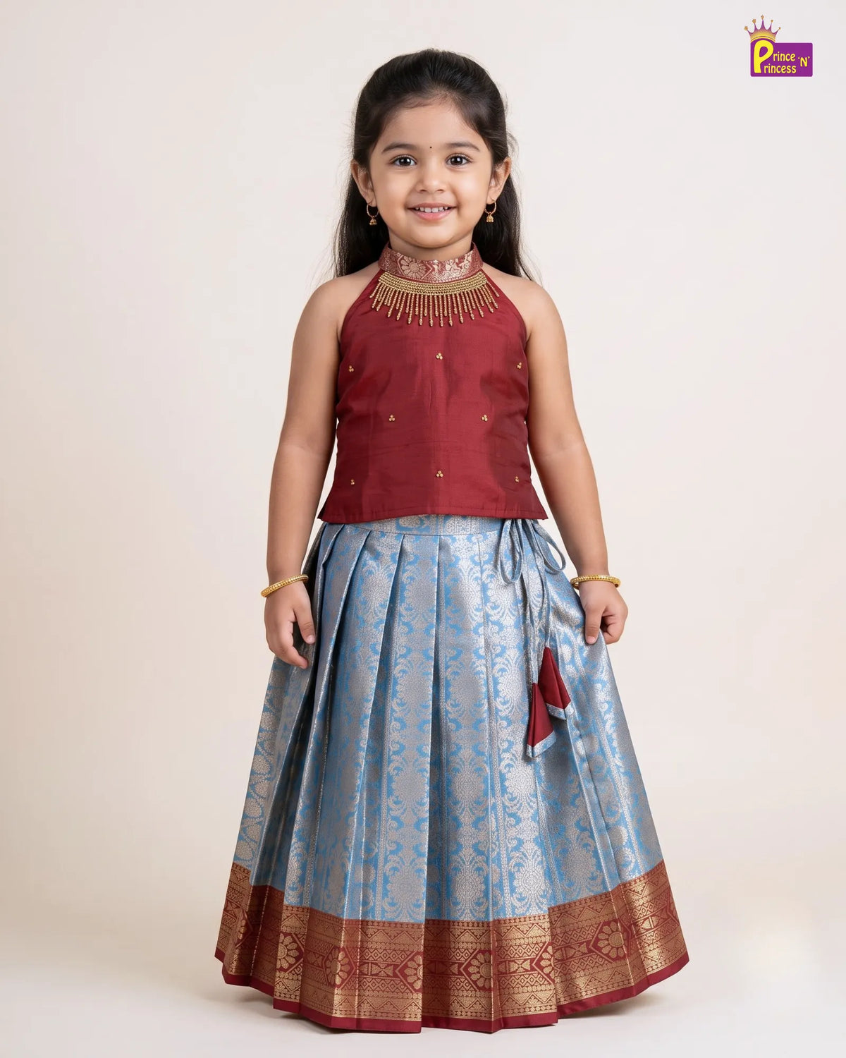 A young girl modeling a traditional Indian outfit consisting of a maroon sleeveless top with gold embroidery and a light blue patterned skirt with a gold border, set against a plain beige background.