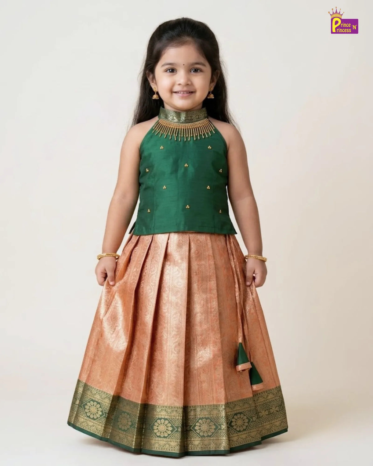 A young girl modeling a traditional Indian ethnic wear set consisting of a sleeveless dark green top with gold embroidery and a pleated peach-beige skirt with a green and gold zari border, against a plain cream background.