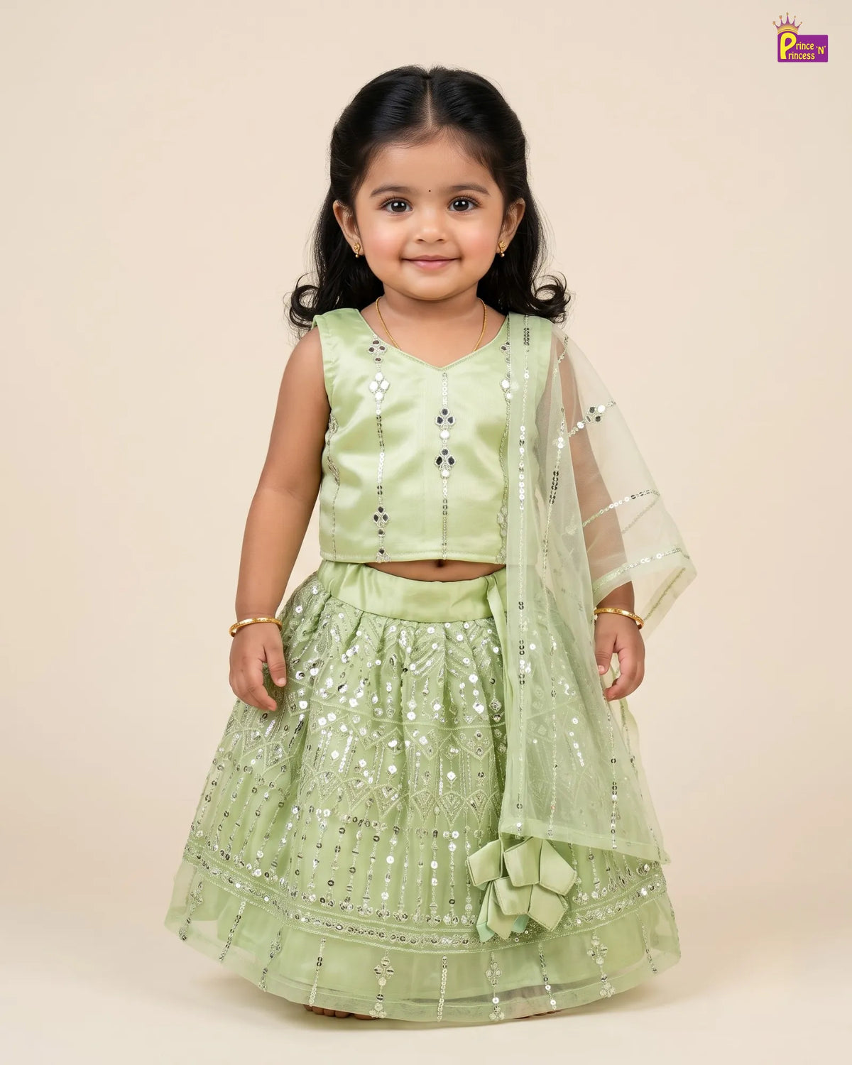 A young girl modeling a light green embroidered traditional Indian outfit with a sleeveless crop top, flared embellished skirt, and a matching sheer dupatta, set against a solid beige background.