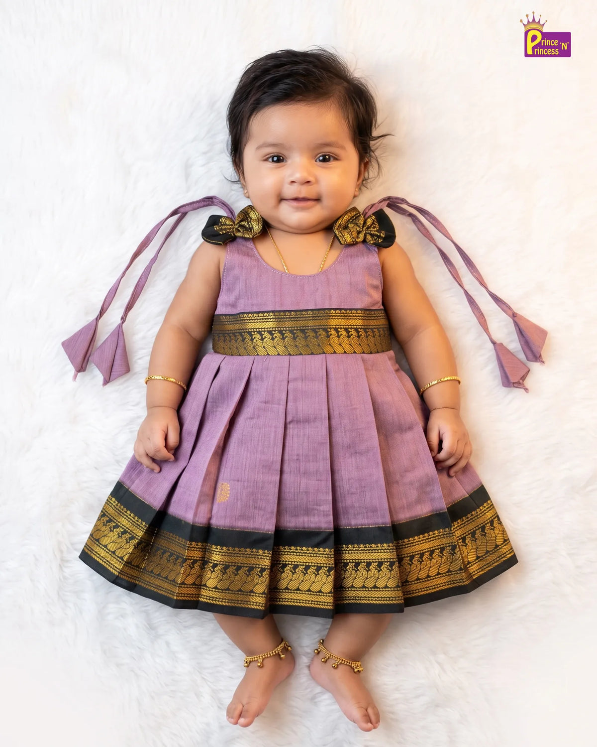 A baby girl wearing a lavender purple traditional dress with gold and black borders, lying on a white textured rug. The dress has decorative bows on the shoulders with purple and gold fabric, and the baby is wearing gold ankle bracelets.