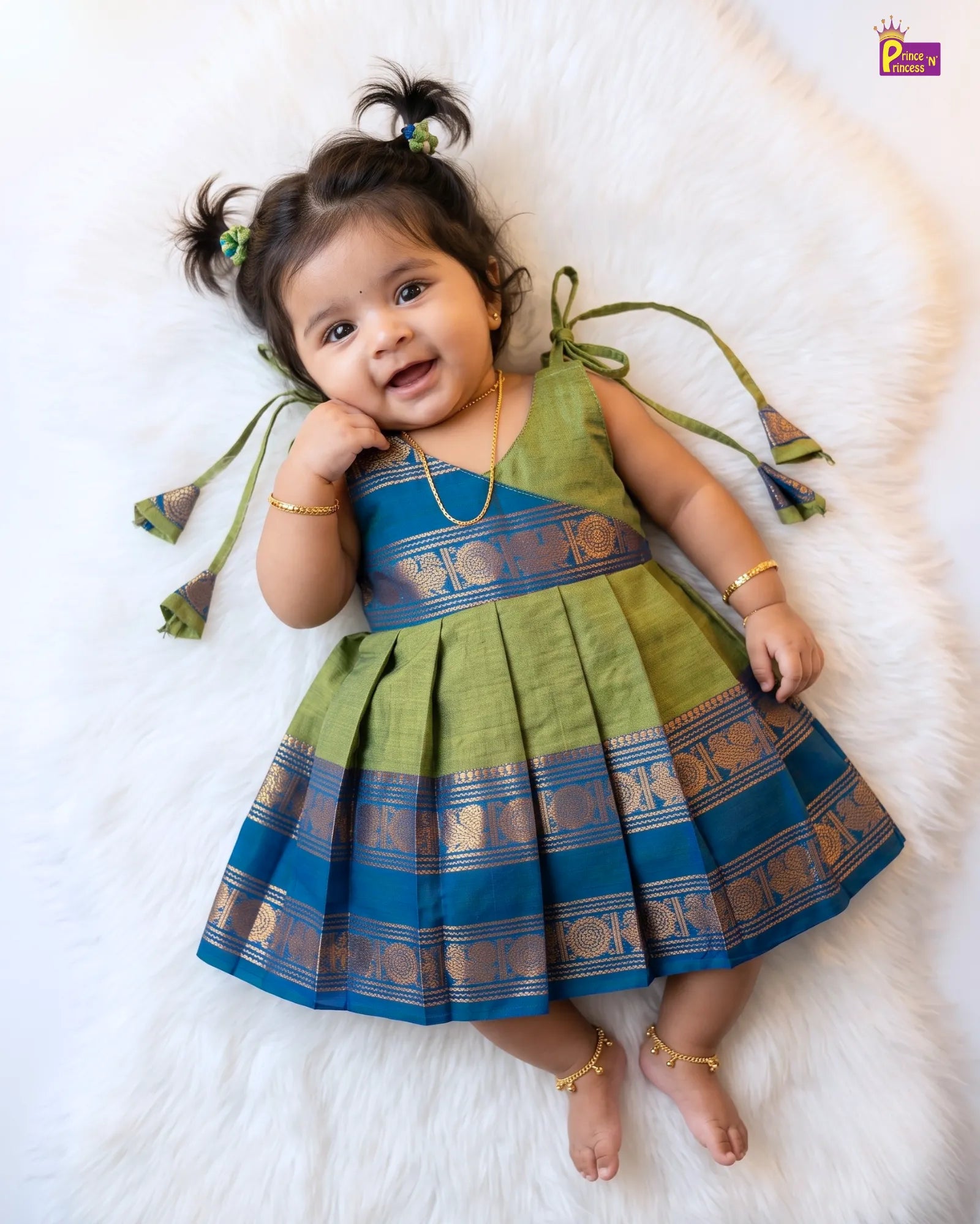 A toddler girl lying on a white faux fur rug wearing a green and blue traditional Indian dress with gold zari borders. The dress has tie-shoulder straps and a pleated skirt.