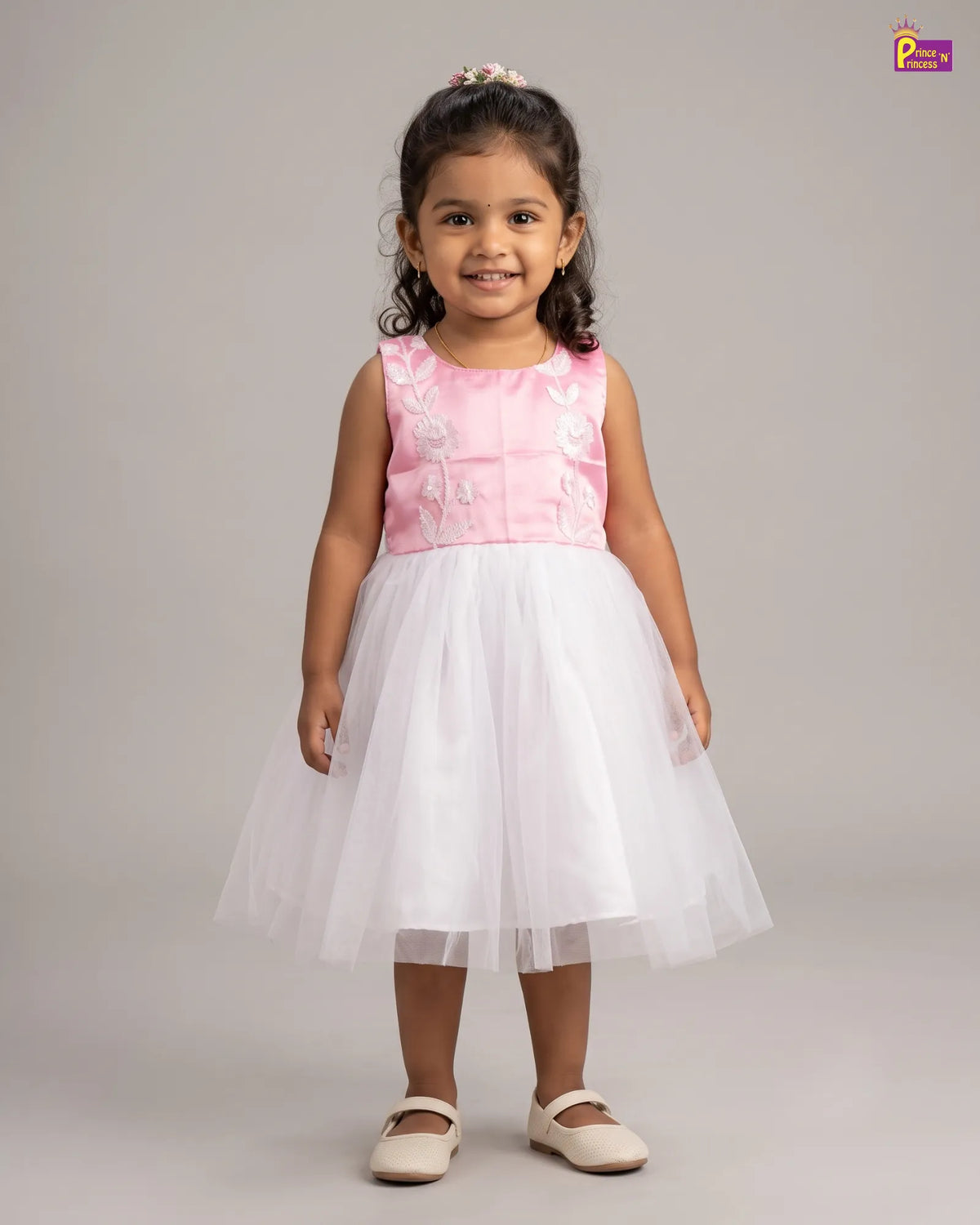 A young girl stands against a light gray studio background wearing a sleeveless pink and white dress with a white tulle skirt and white ballet flats.