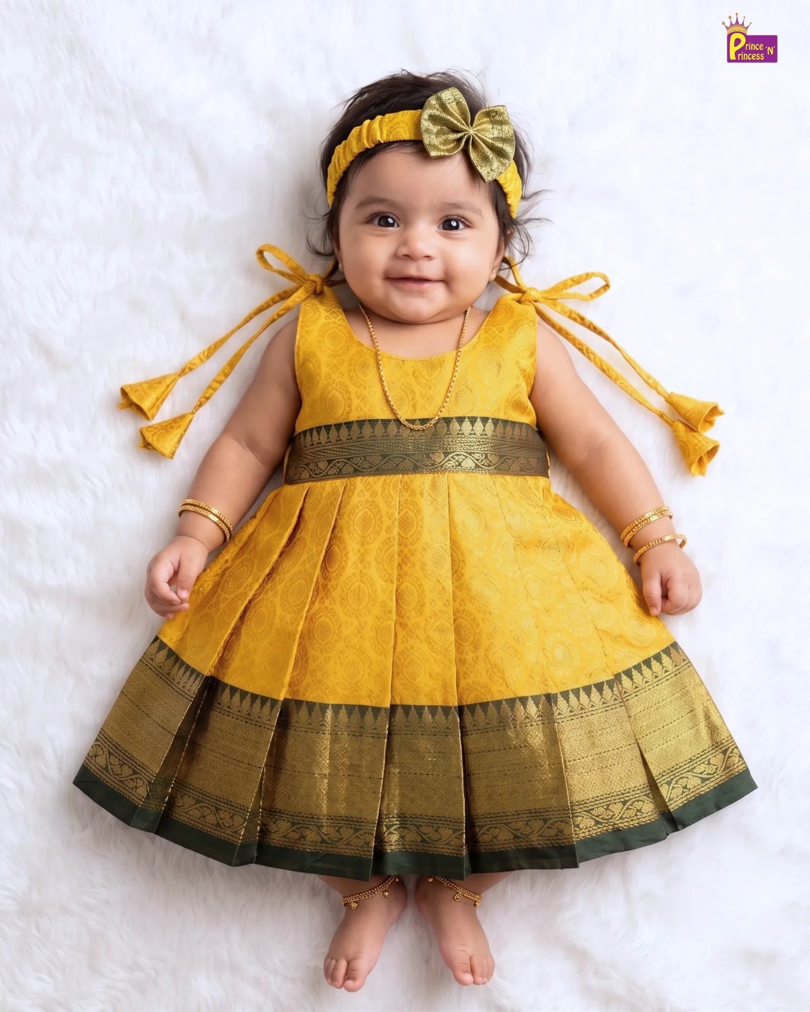 A baby girl wearing a yellow and gold traditional Indian dress with a matching headband, lying on a white textured blanket.