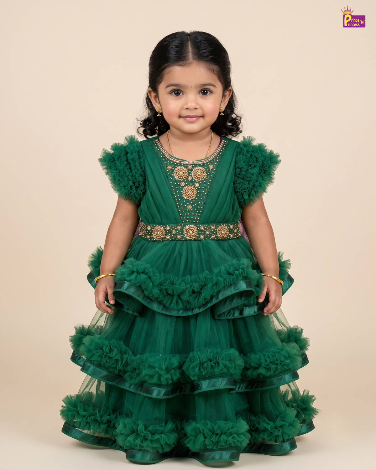 A young toddler girl wearing a green tiered tulle dress with gold embroidery and ruffled tulle details, standing against a solid beige background.