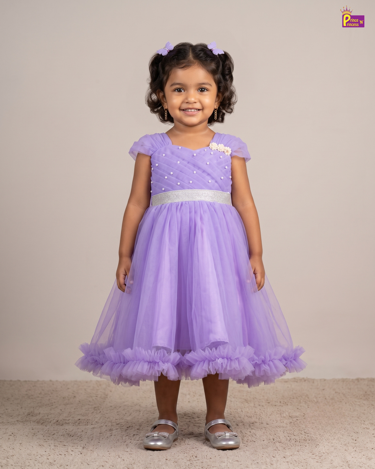 A young girl with curly dark hair wearing a lavender purple tulle party dress with a silver sash and silver shoes, standing against a plain beige studio background.