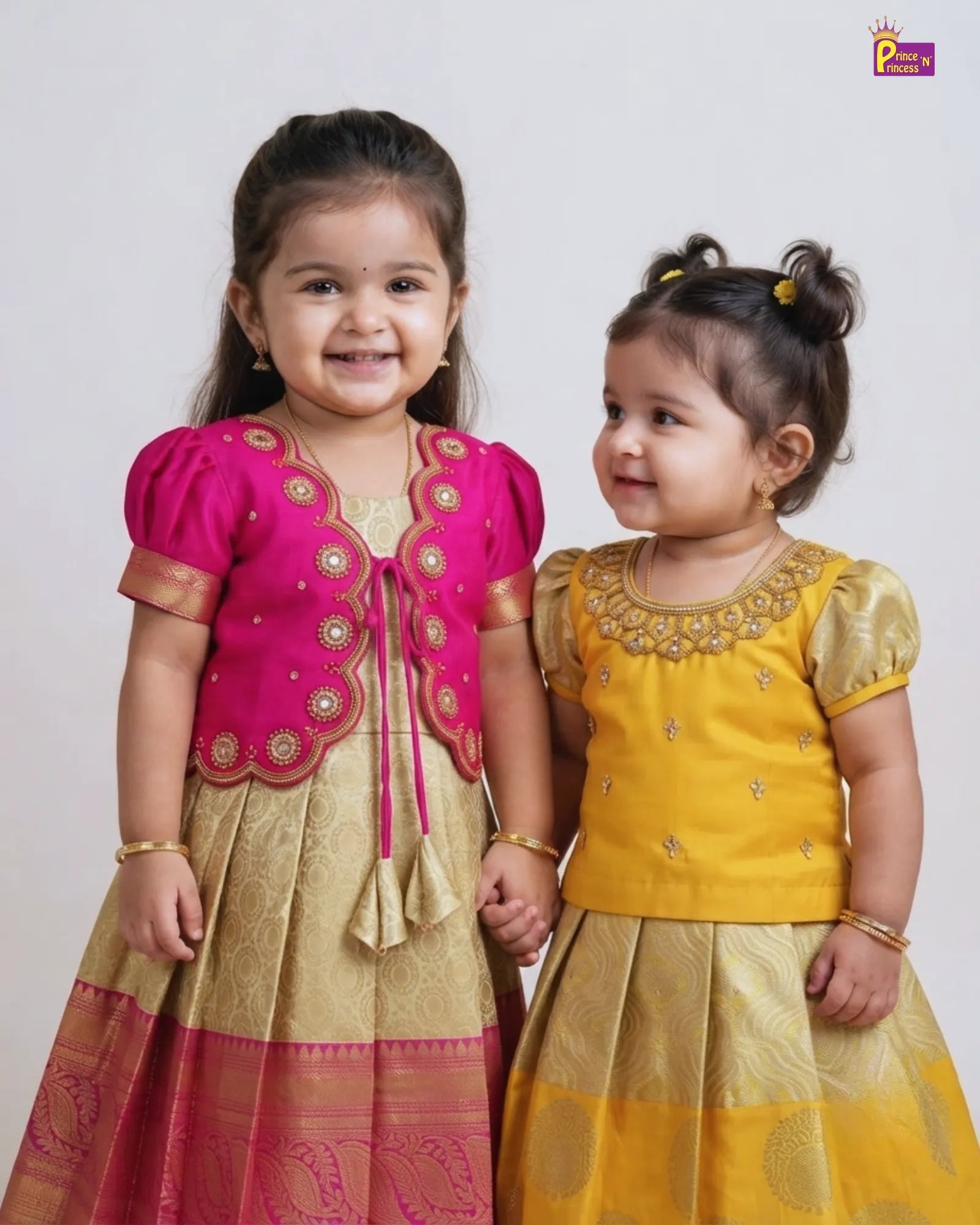 Two young girls wearing traditional Indian ethnic wear, standing side-by-side and holding hands. The girl on the left wears a bright pink embroidered kurta with a gold-patterned skirt, and the girl on the right wears a mustard yellow embroidered kurta with a matching skirt.
