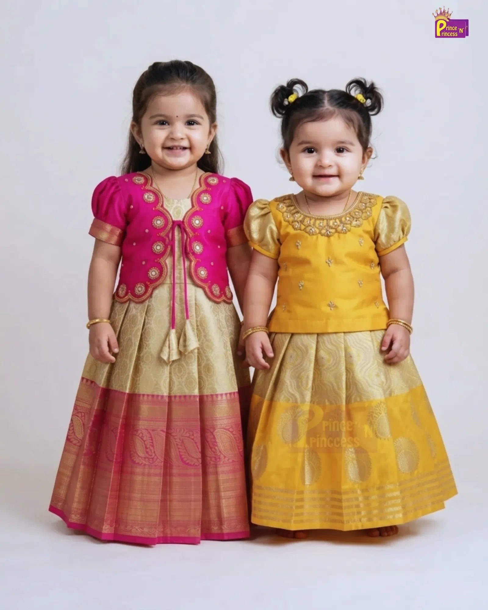Two young girls modeling traditional Indian ethnic wear, standing side-by-side against a light gray studio background. The girl on the left wears a pink and gold embroidered kurta and skirt set, while the girl on the right wears a yellow and gold embroidered kurta and skirt set.