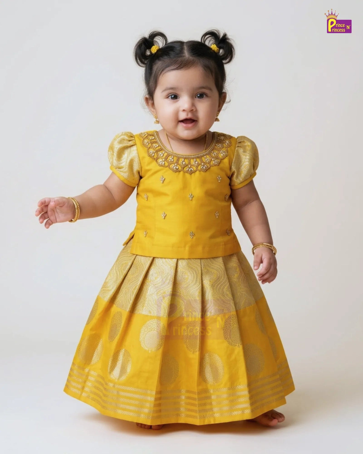 A toddler girl wearing a traditional Indian yellow dress with gold embroidery and a pleated skirt, standing against a plain light background.