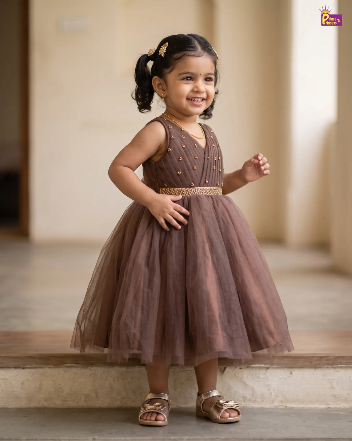 A young girl with dark hair in a ponytail stands smiling on outdoor steps, wearing a sleeveless brown tulle dress with gold embellishments and sandals.