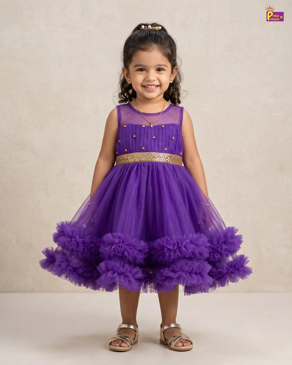 A young girl wearing a sleeveless purple tulle dress with a gold embellished bodice, a gold waistband, and a voluminous ruffled tulle skirt, standing against a neutral beige studio backdrop.