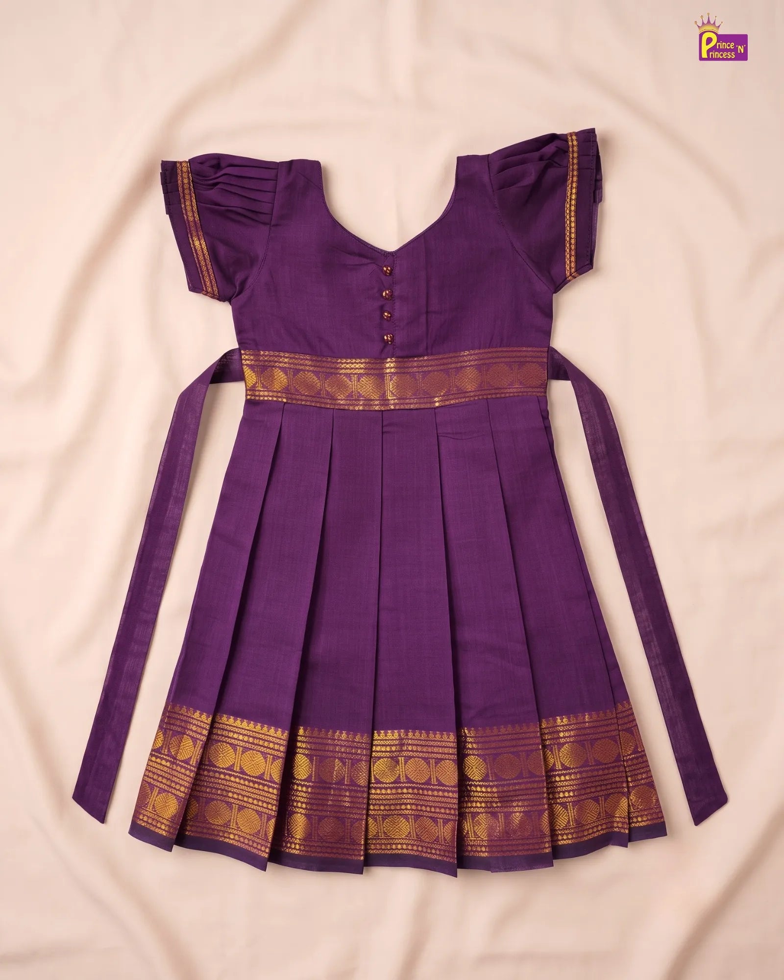 A purple traditional Indian dress with gold zari borders, laid flat on a light beige fabric background. The dress features short flutter sleeves, a V-neckline with decorative buttons, a pleated skirt, and long side ties.