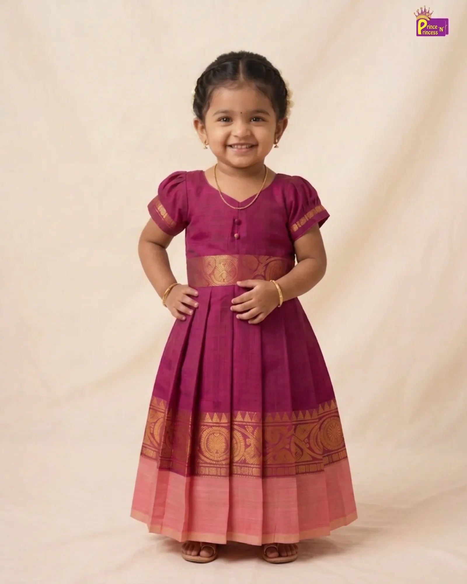 A young girl modeling a traditional Indian salwar kameez dress in magenta and pink with gold zari embroidery, standing against a neutral beige backdrop.