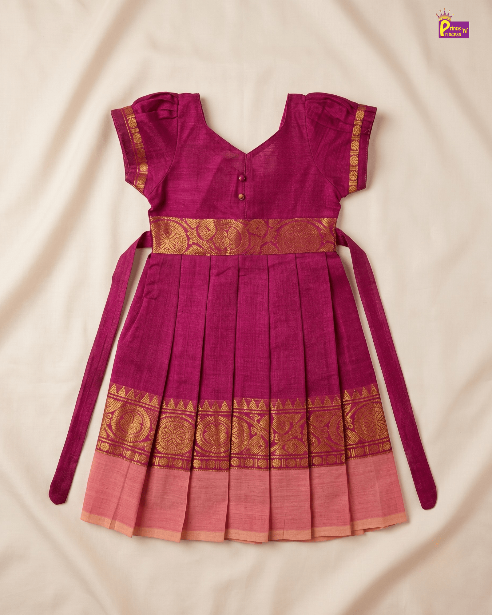 A magenta pink traditional Indian dress with gold zari embroidery, featuring a pleated skirt, short sleeves, and a waist tie, laid flat on a cream-colored fabric background.