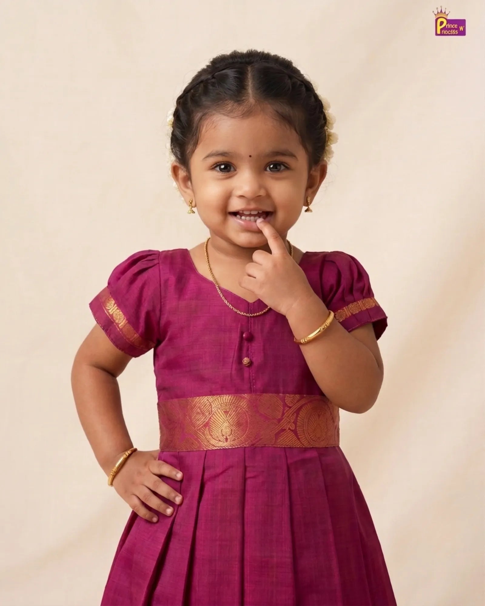 A young girl with dark hair in a braided style, wearing a magenta traditional Indian dress with gold zari embroidery and a gold waistband, standing against a plain beige background.