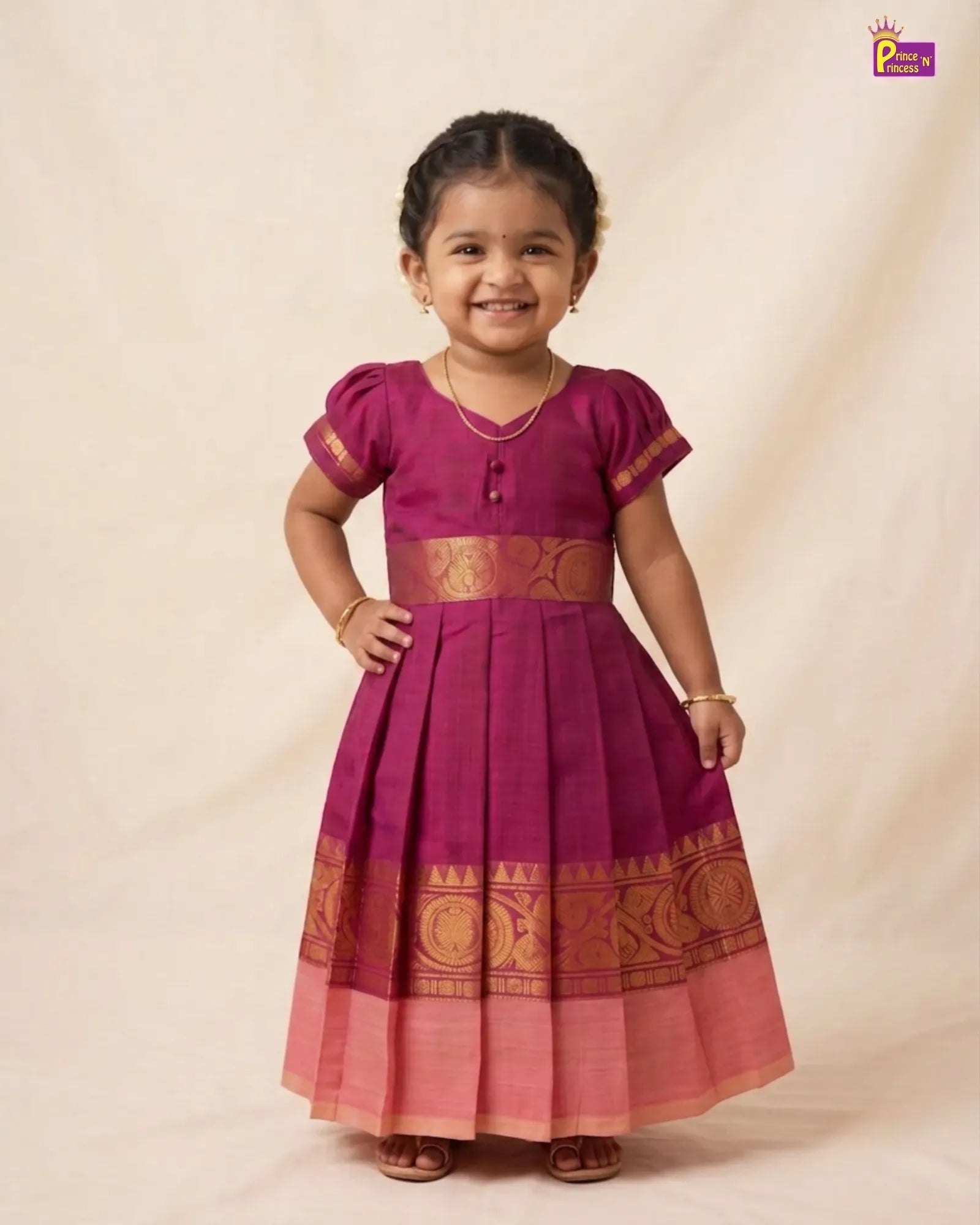 A young girl smiling and wearing a magenta purple and pink traditional Indian dress with gold zari embroidery, standing against a beige fabric backdrop.