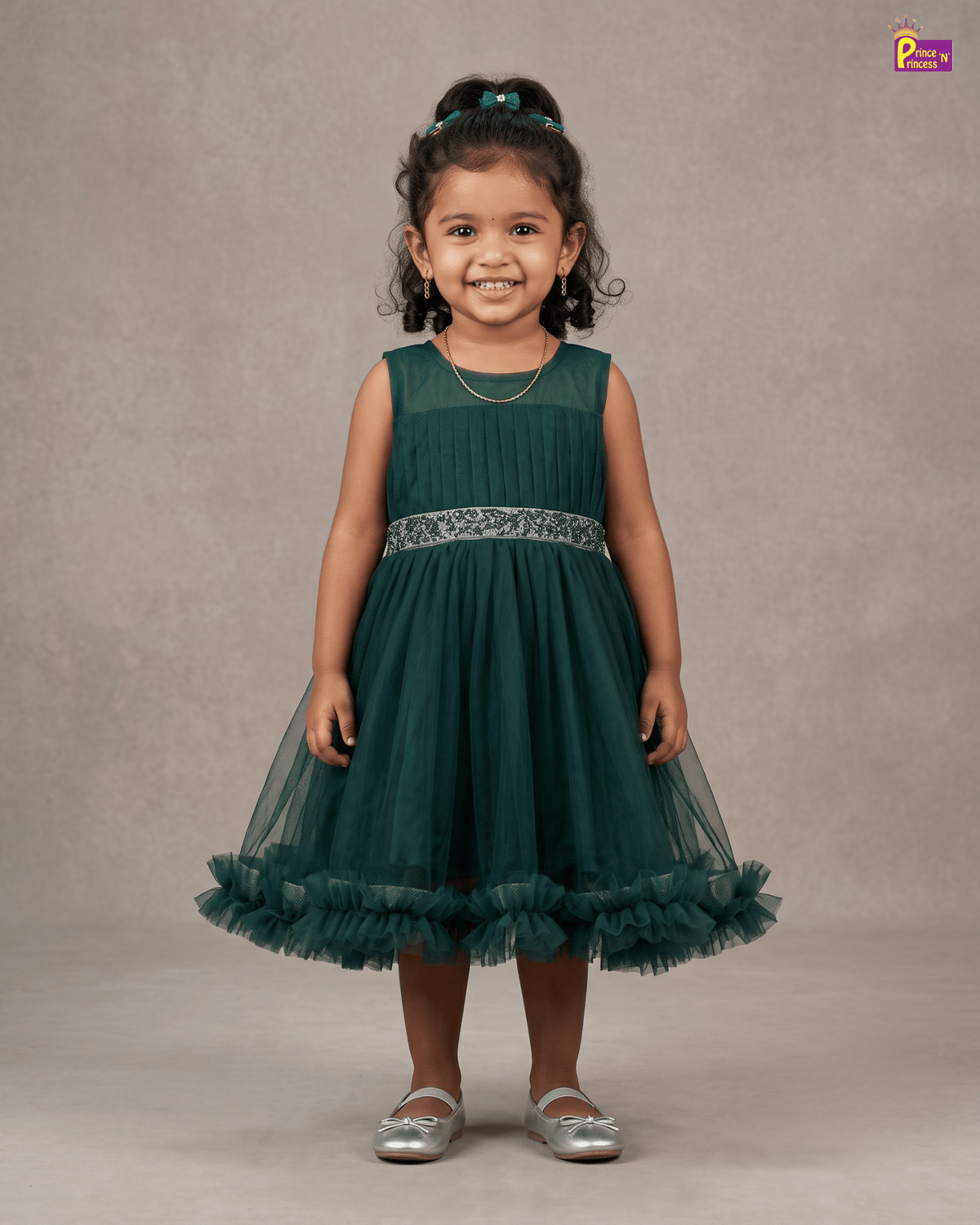 A young girl with curly dark hair wearing a sleeveless dark green tulle flower girl dress with a ruffled hem and a silver patterned waistband, standing against a neutral gray studio backdrop.