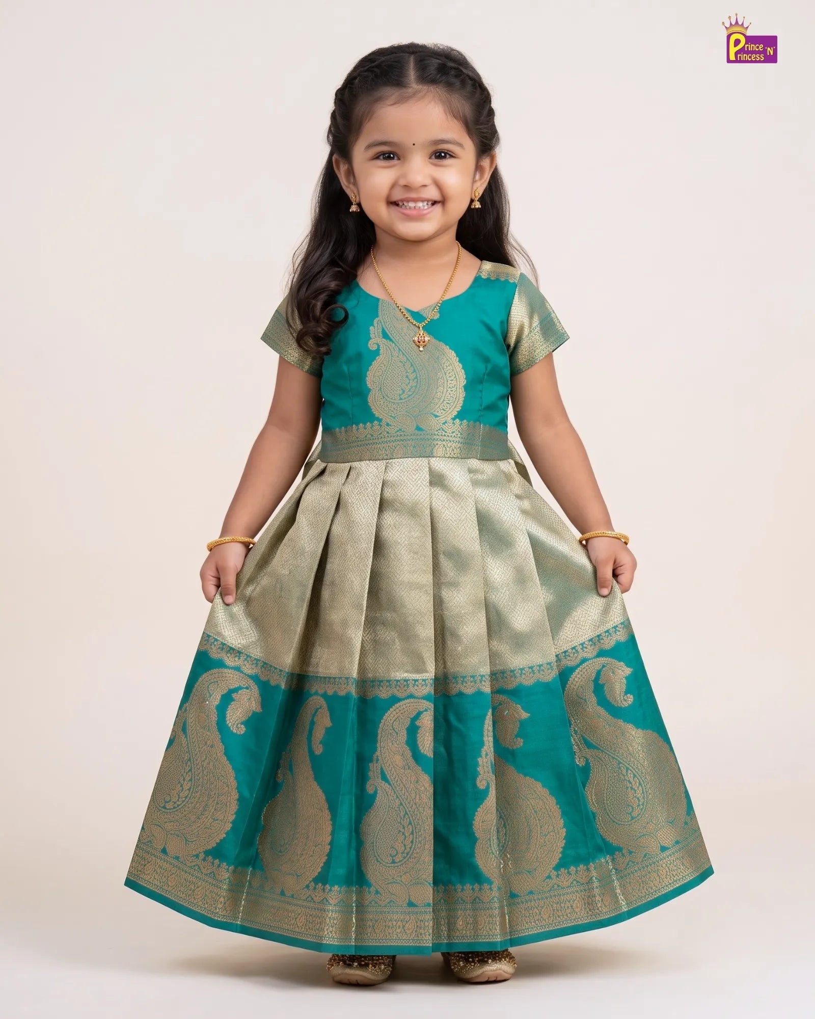 A young girl wearing a traditional Indian salwar kameez dress in teal green and beige with gold zari embroidery, standing against a plain light beige background.