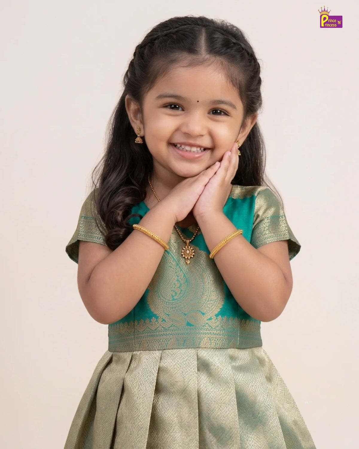 A young girl smiling and posing in a traditional Indian outfit with a teal green and gold embroidered top and a light gold skirt, set against a plain light beige background.