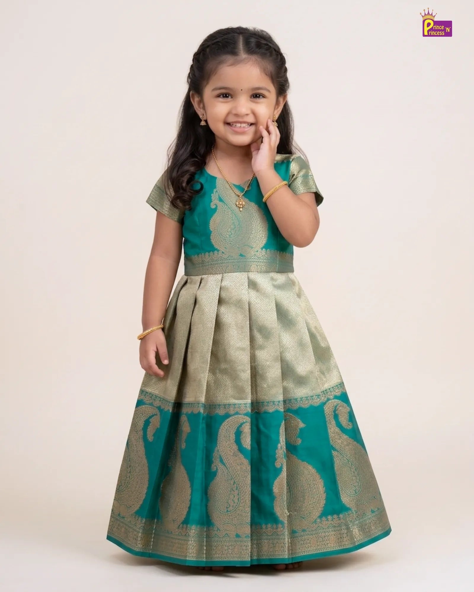 A young girl modeling a traditional Indian Anarkali gown in teal green and gold zari. The dress features a short-sleeved bodice with a floral pattern, a pleated skirt, and a wide border with paisley motifs.