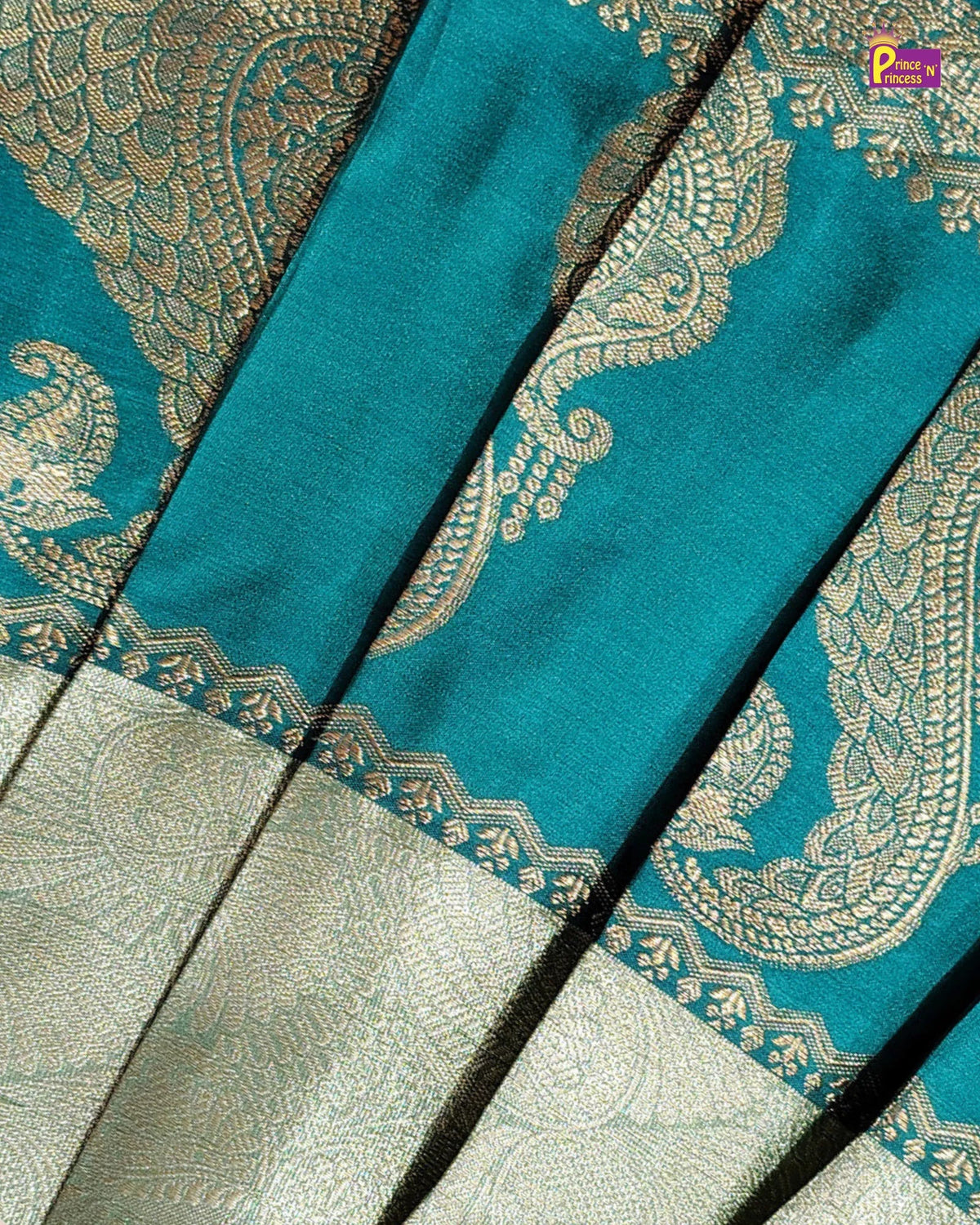 A close-up of a teal blue fabric with intricate gold paisley and floral brocade patterns, layered over a light beige patterned textile. A small 'P Pindora' brand logo is visible in the top right corner.