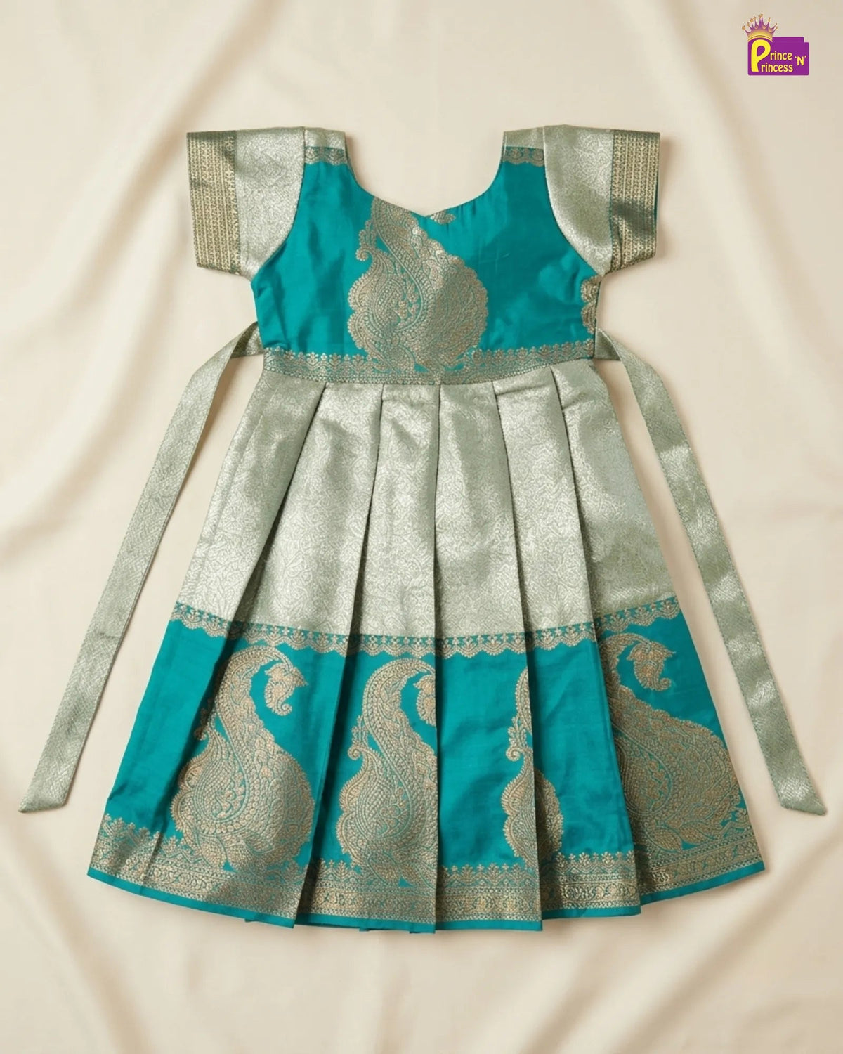 A traditional Indian girl's dress (Anarkali) in teal and silver brocade fabric, laid flat on a cream-colored fabric background. The dress features a teal bodice with silver paisley motifs, silver sleeve borders, a pleated silver skirt, and long tie-garlands.