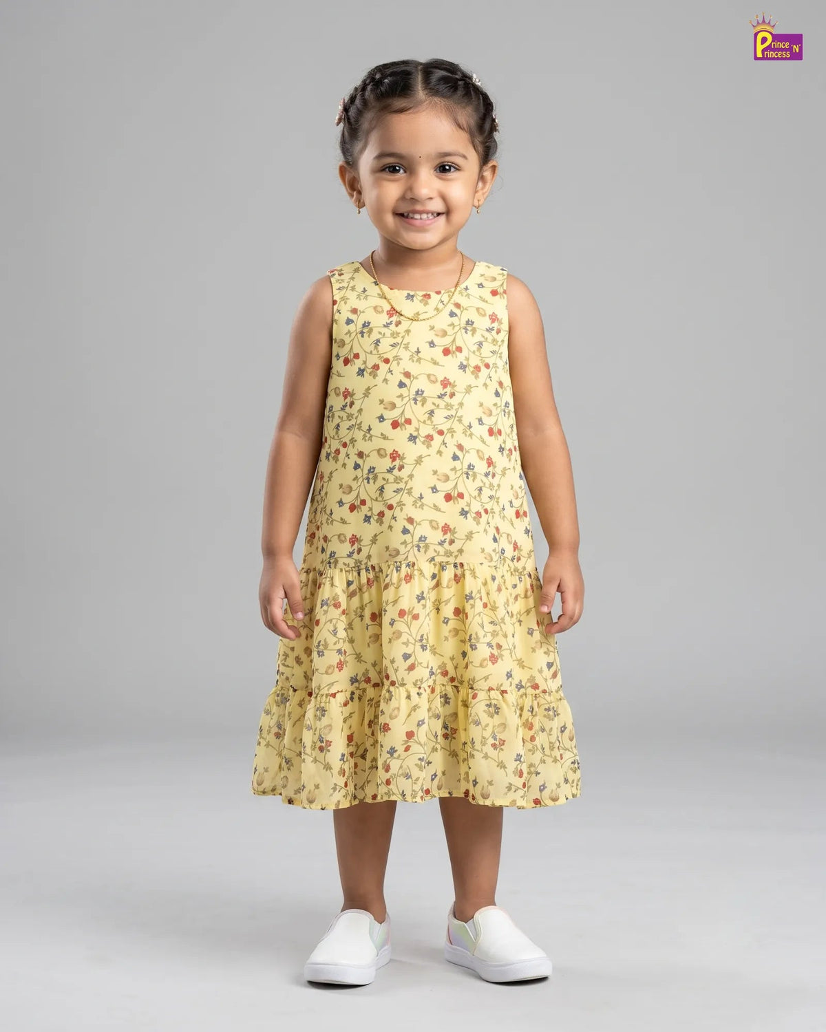 A young girl with braided hair stands against a light gray studio background wearing a sleeveless tiered yellow dress with a small multicolor floral print and white slip-on shoes.