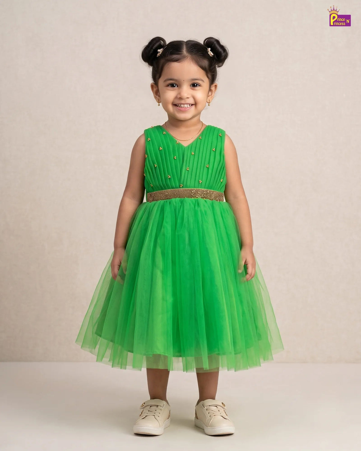 A young girl with her hair in two buns stands wearing a sleeveless bright green party dress with a pleated bodice, a glittery gold waistband, and a layered tulle skirt. She is wearing off-white sneakers against a neutral beige studio background.