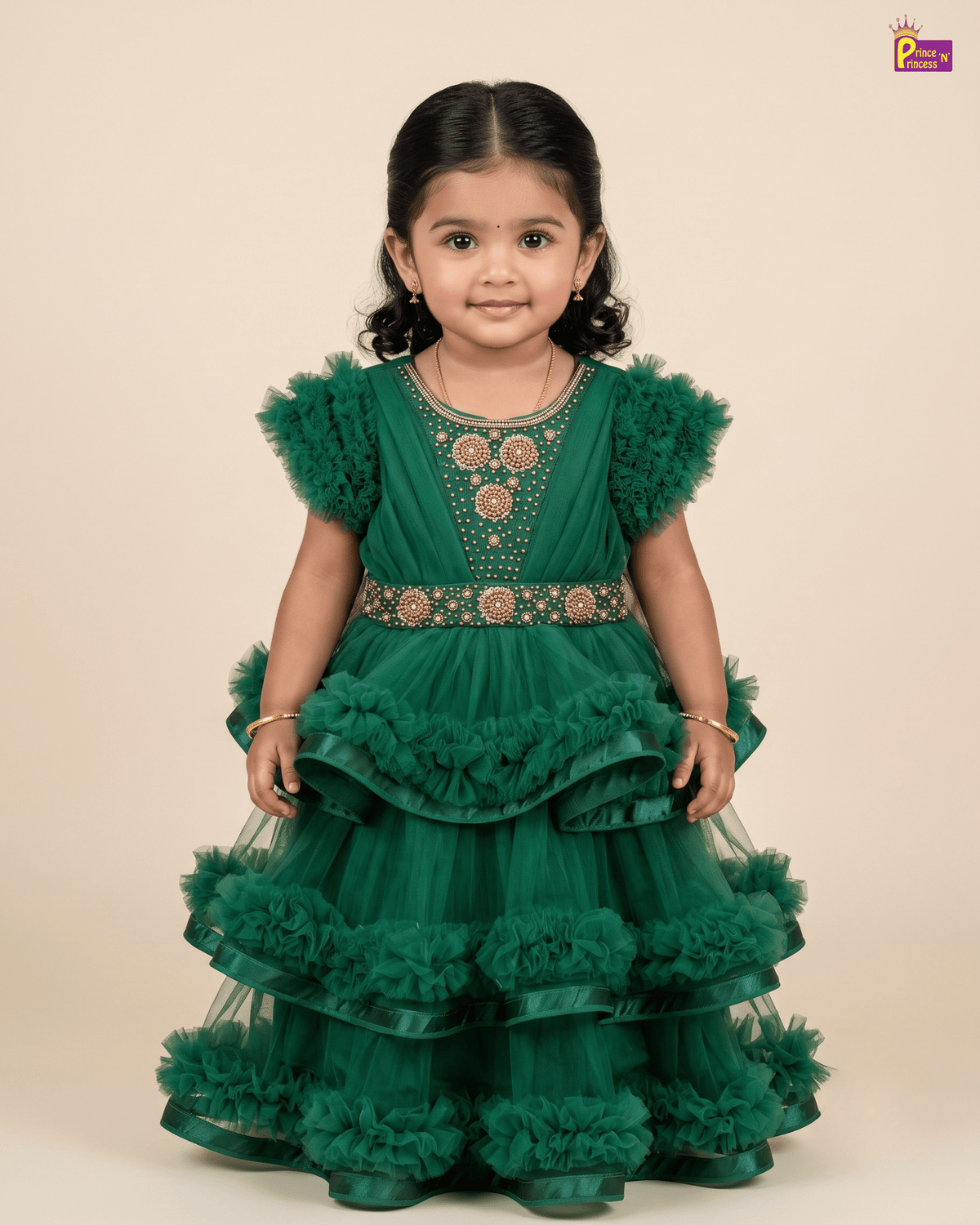 A young toddler girl wearing a green tiered tulle dress with gold embroidery and ruffled tulle details, standing against a solid beige background.