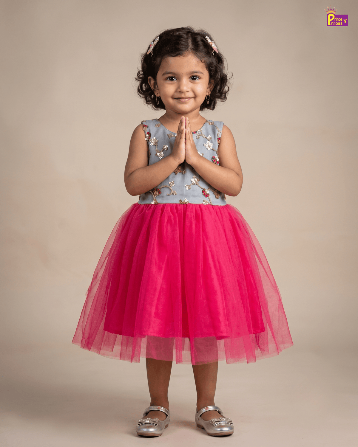 A young girl with curly dark hair wearing a sleeveless light blue floral top and a bright pink tulle skirt, standing with hands pressed together in a studio setting.