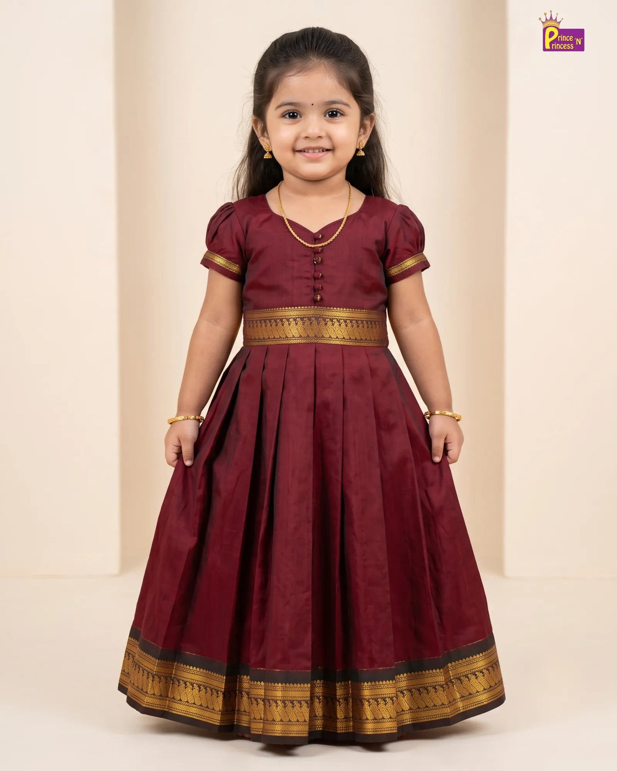 A young girl modeling a maroon A-line dress with gold zari borders and button details on the bodice, standing against a neutral beige background.