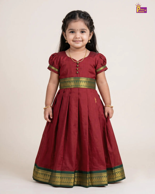 A young girl modeling a traditional Indian maroon red Anarkali gown with gold and green zari borders, standing against a plain beige studio background.