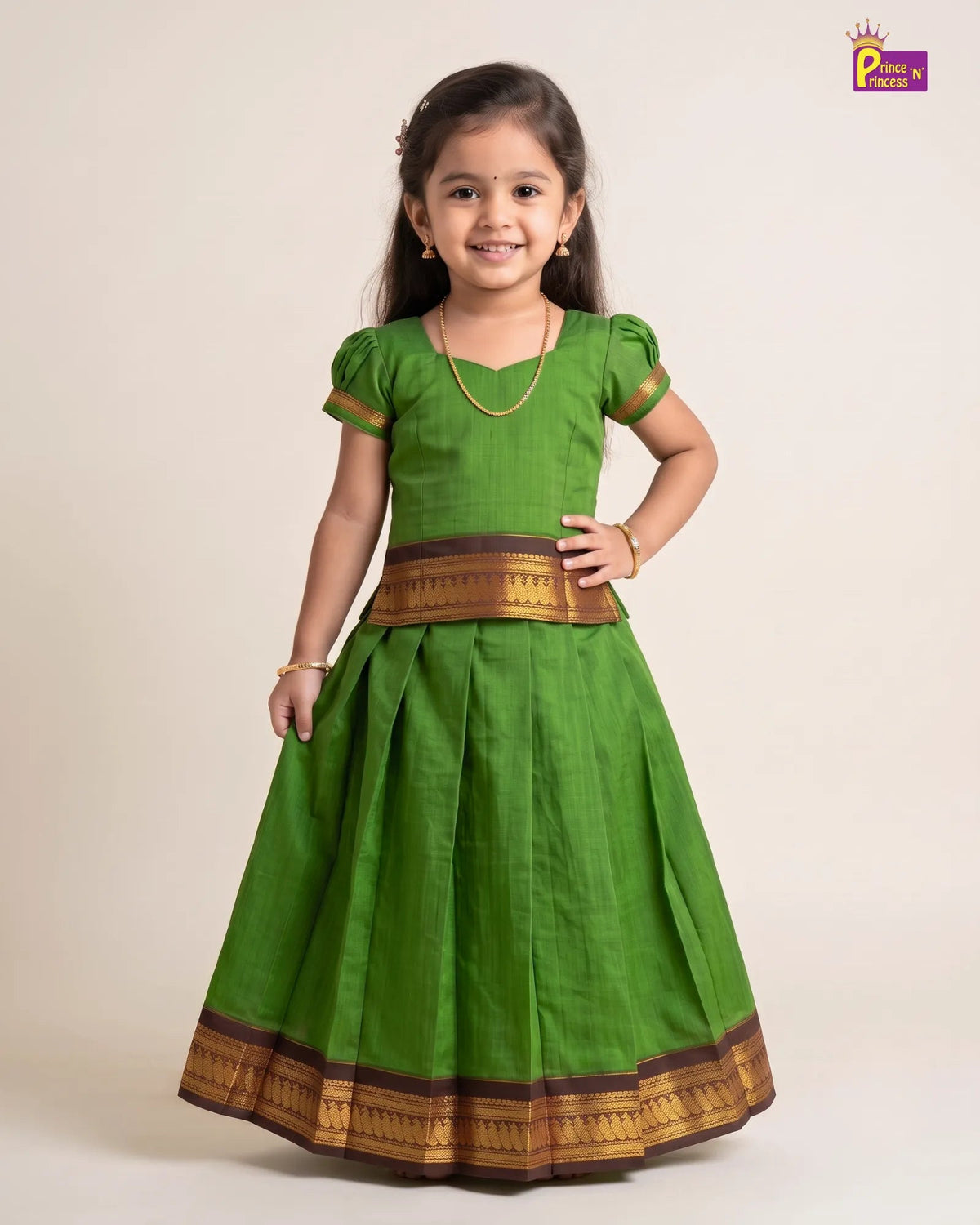 A young girl modeling a green traditional Indian dress with gold and brown borders, standing against a plain beige studio background.