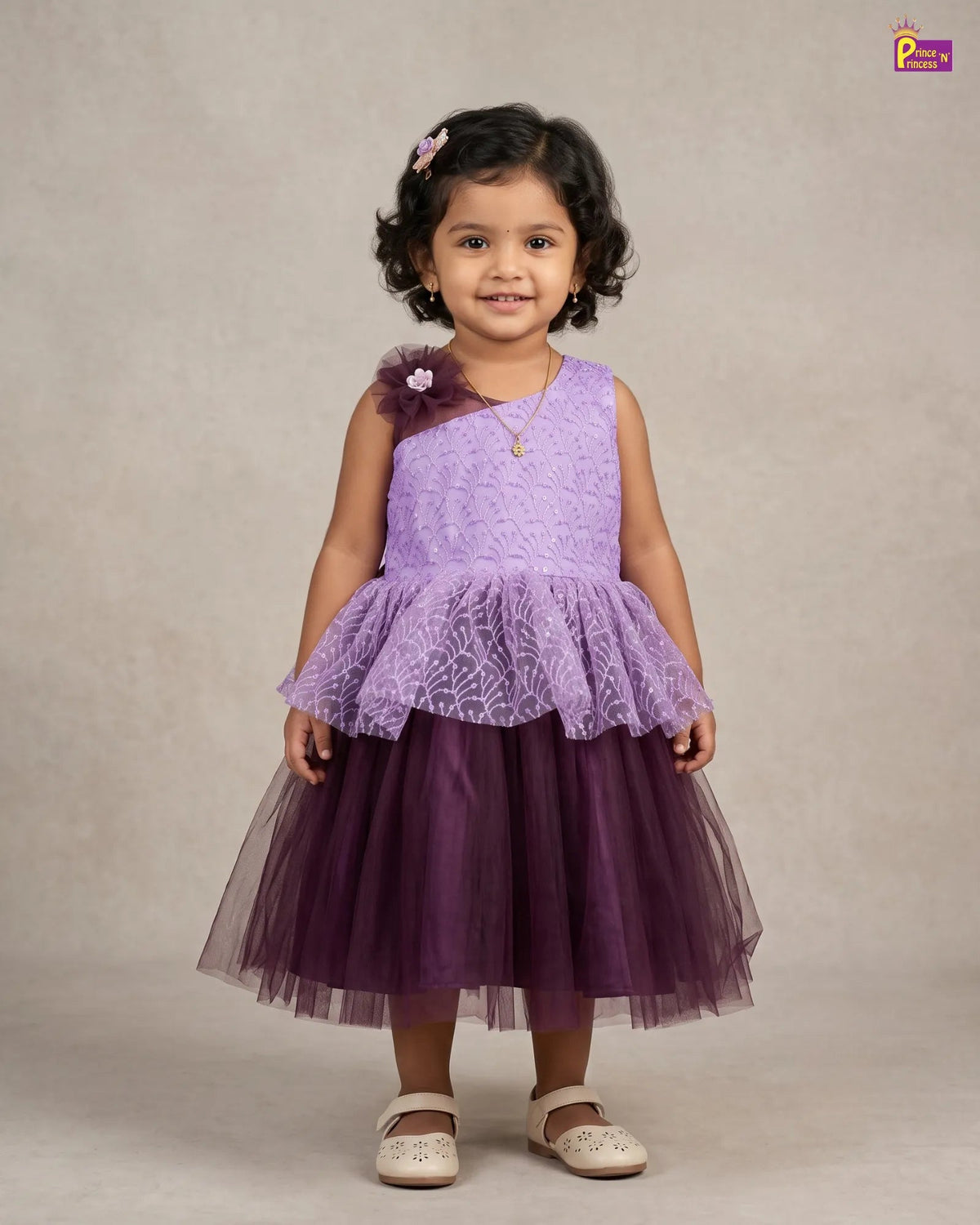 A young girl with dark curly hair wearing a two-tone purple dress, standing against a neutral beige studio background. The dress features a light purple lace one-shoulder top with a ruffled overlay and a darker purple tulle skirt.