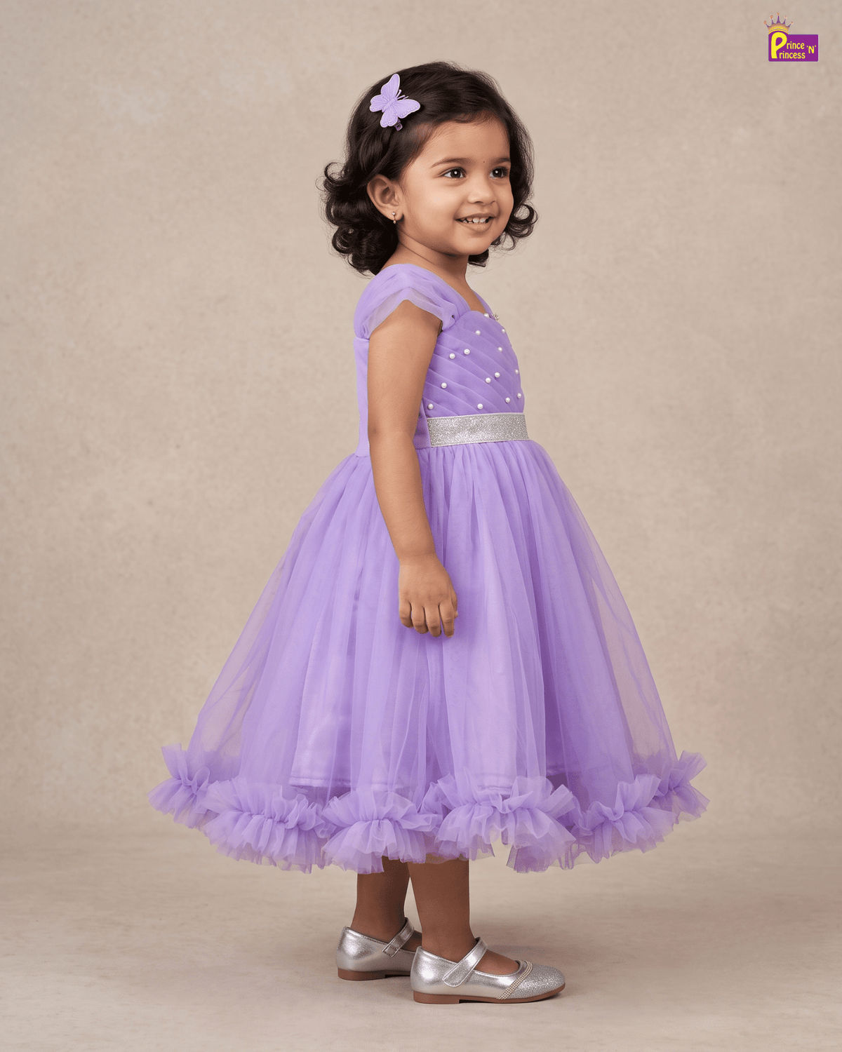 A young girl with dark curly hair wearing a lavender purple party dress with a sparkly bodice, silver belt, and ruffled tulle skirt, standing against a beige studio backdrop.